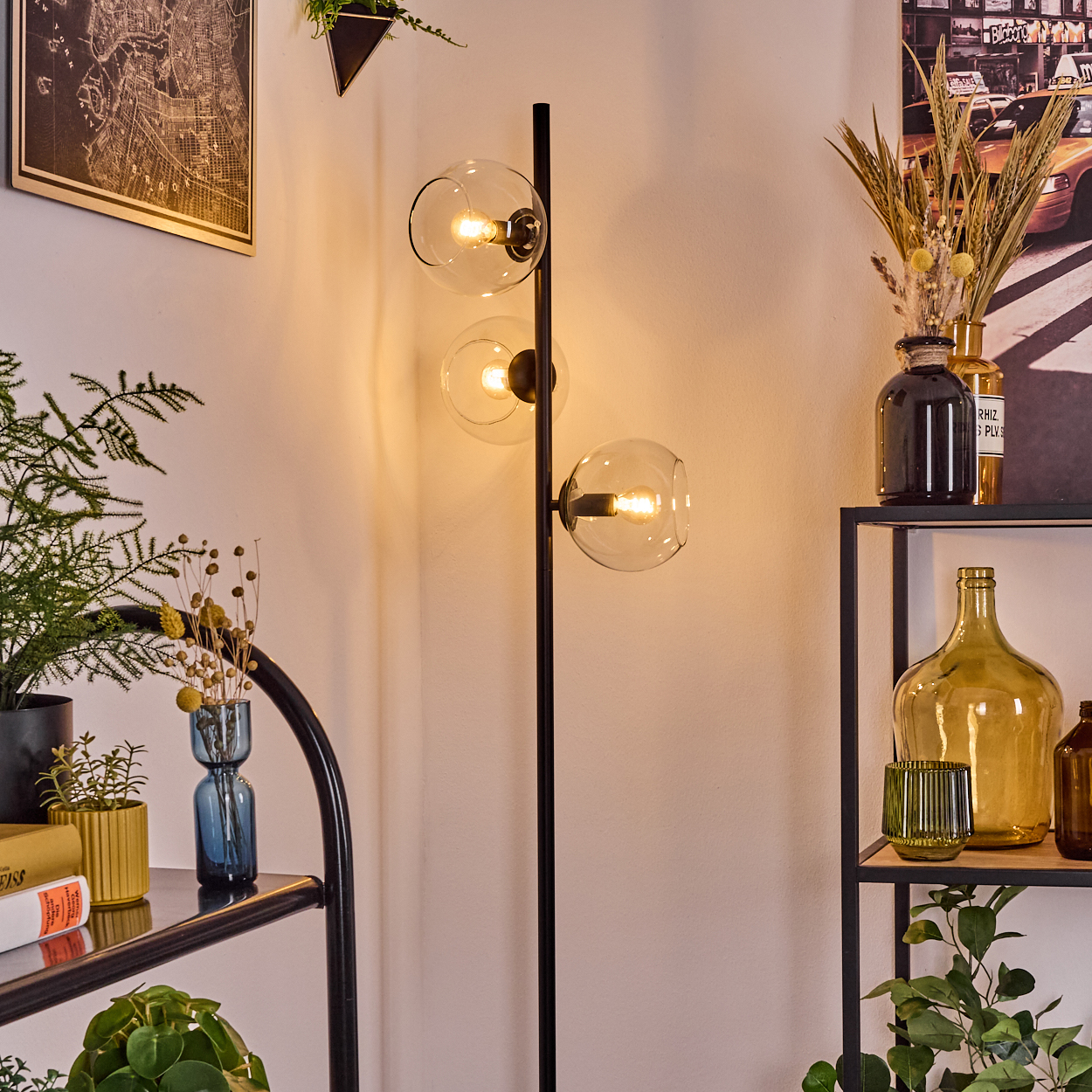 Eine moderne Stehlampe mit drei Glaskugeln. Regale halten Vasen, Pflanzen und Bücher. Eine Karte und ein Bild an der Wand.