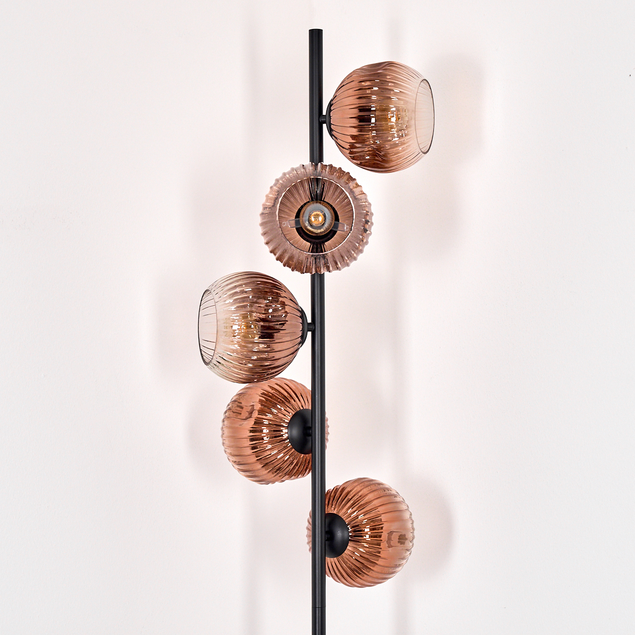 Eine moderne Stehlampe mit fünf kupferfarbenen Glasschirmen, die vertikal an einer schwarzen Stange angeordnet sind, vor einer weißen Wand.