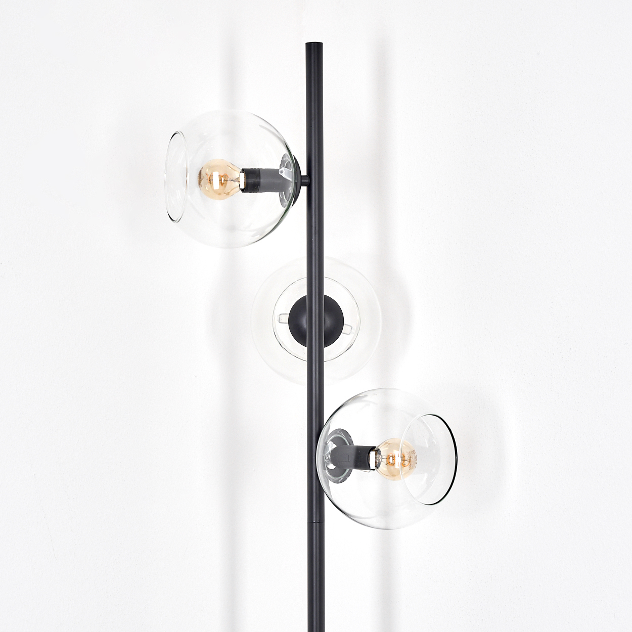 Eine moderne schwarze Stehlampe mit drei klaren Glaskugelschirmen, jede mit einer warmen Glühbirne, vor einer weißen Wand.