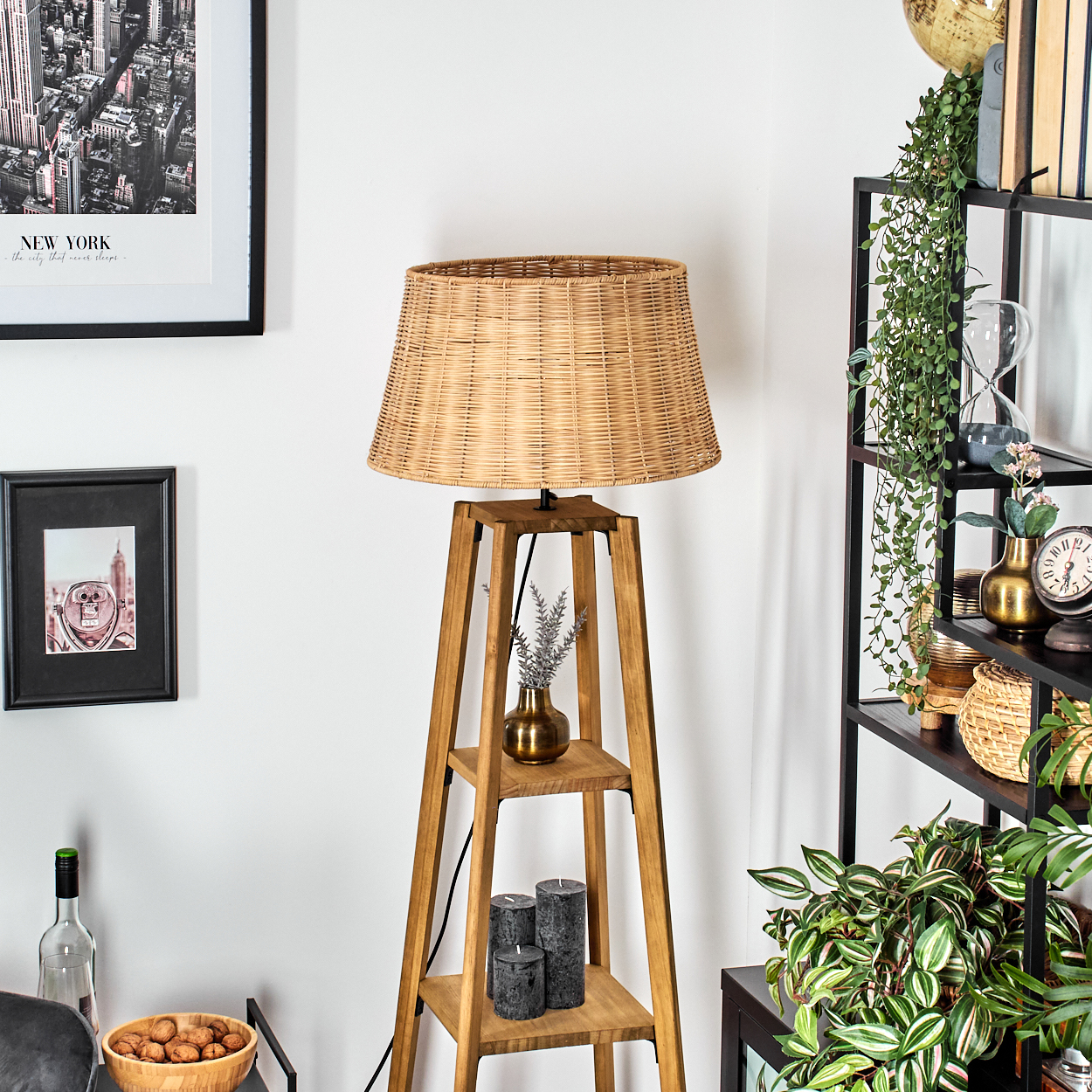 Eine gewebte Lampe steht auf einem Holzständer. Ein Regal hält eine Vase, Kerzen. Ein schwarz gerahmtes Bild und ein New York-Poster hängen an der Wand.