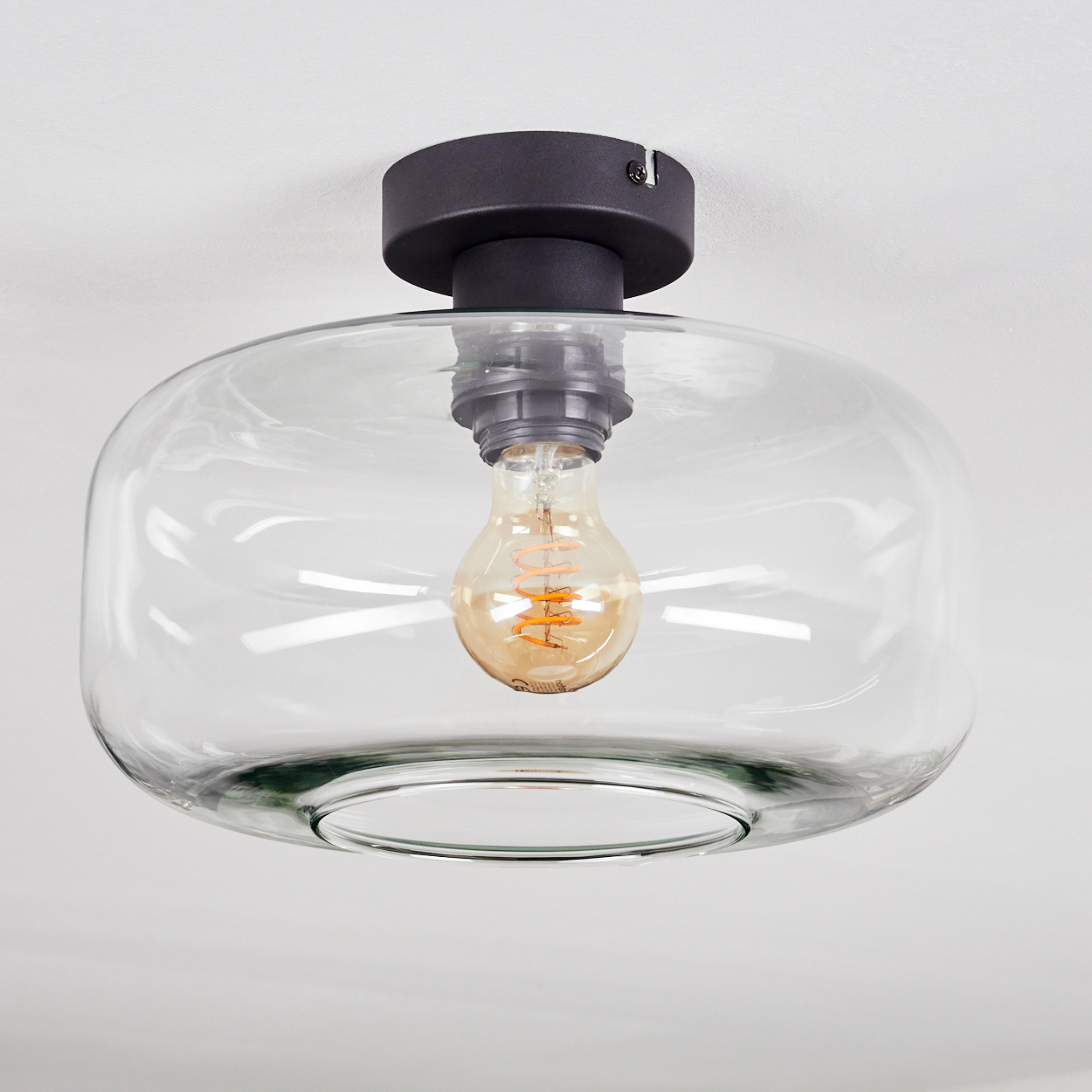 Eine klare Deckenlampe aus Glas mit schwarzem Sockel. Im Inneren befindet sich eine freiliegende Glühbirne. Der Hintergrund ist weiß.