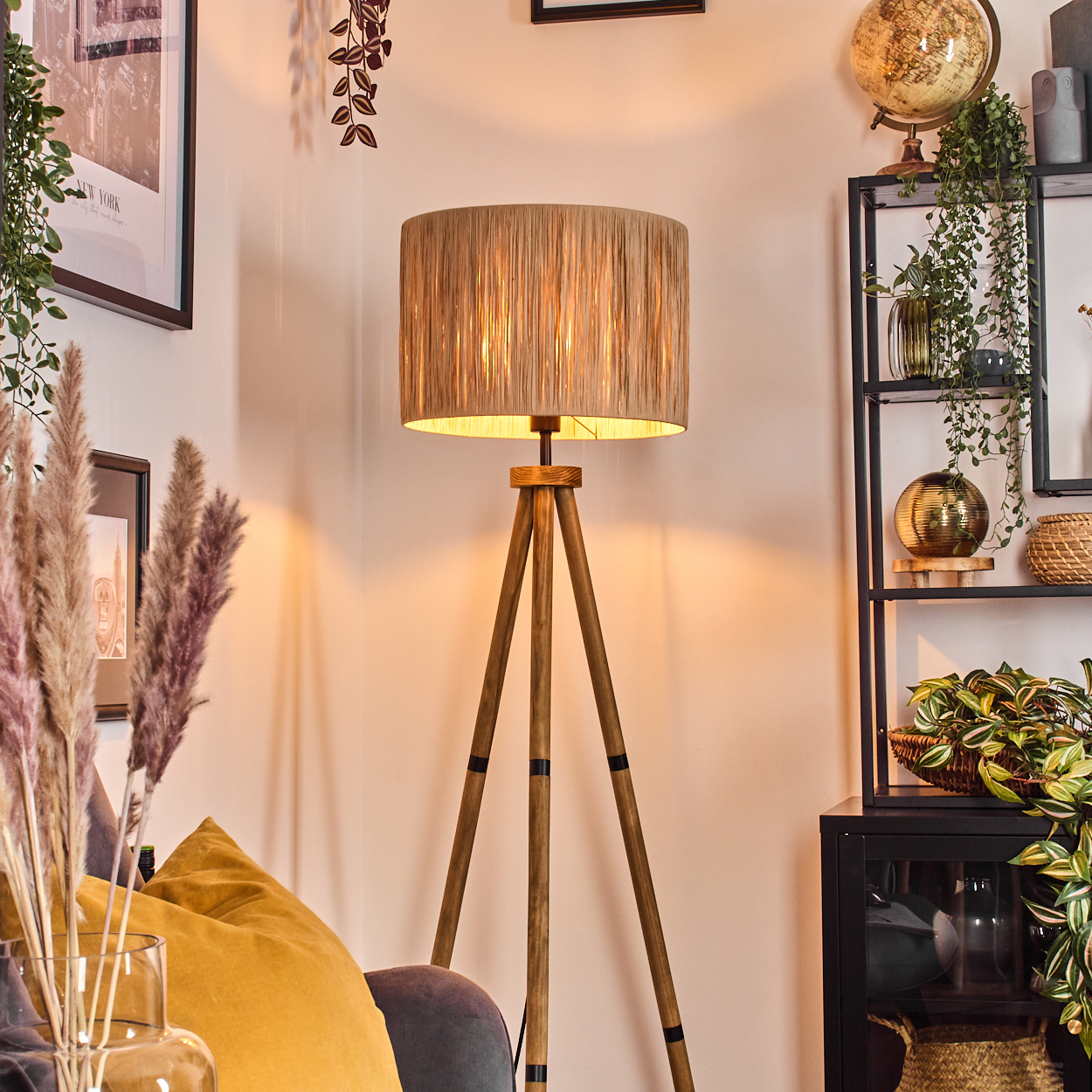 Ein Raum mit einer hölzernen Stativlampe. Ein gelbes Kissen und dekorative Pflanzen stehen vor einem Regal mit Globus und Pflanzen.