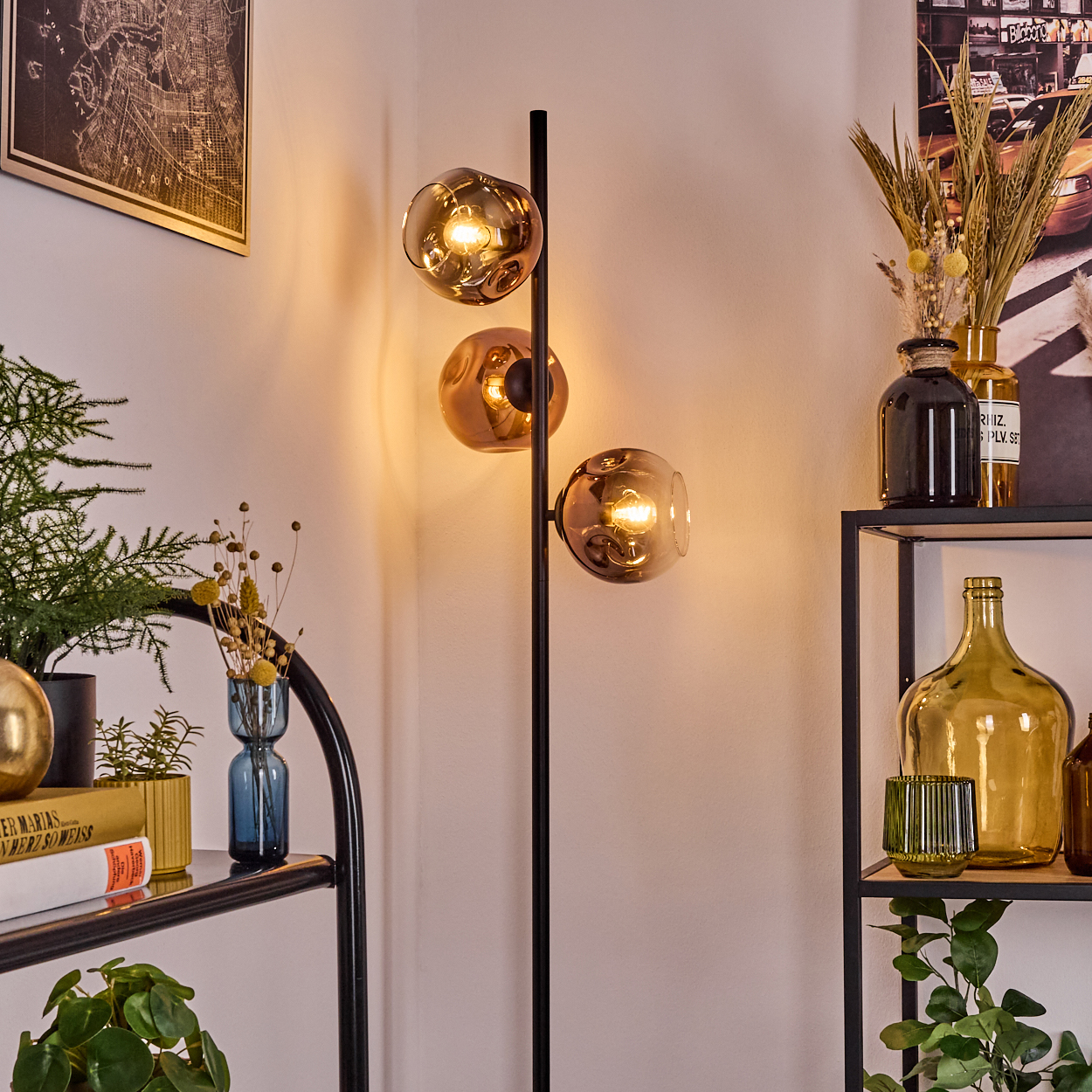 Eine moderne Stehlampe mit drei kugelförmigen, bernsteinfarbenen Glasschirmen. Im Hintergrund sind Regale mit Vasen und Pflanzen zu sehen.