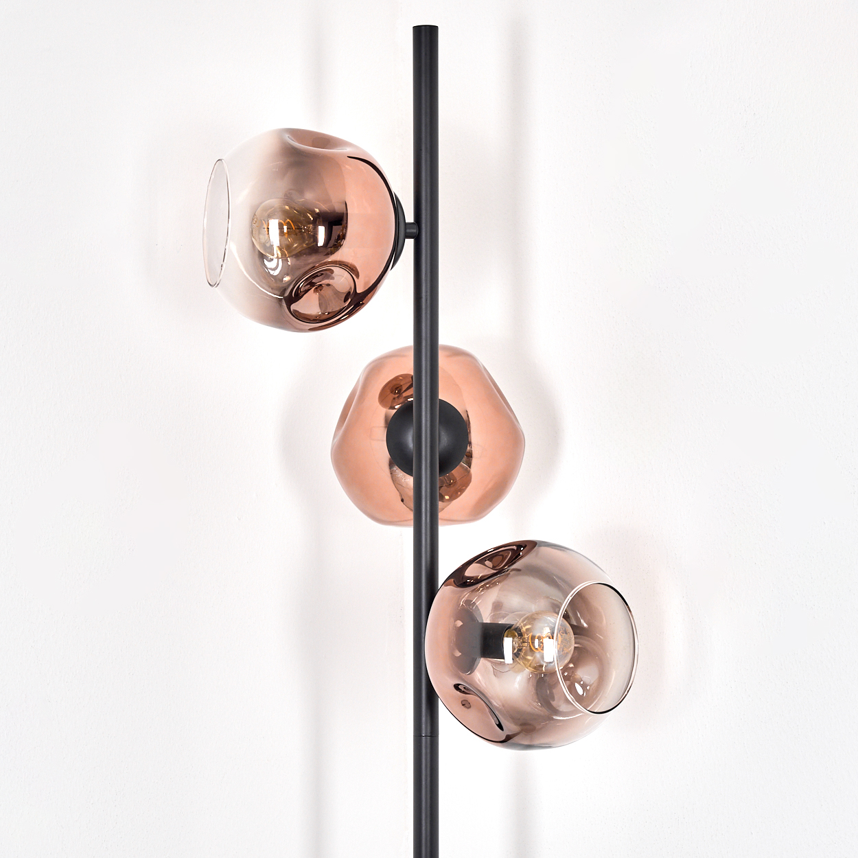 Eine moderne Stehlampe mit drei kugelförmigen, kupferfarbenen Glasschirmen. Die Lampe hat einen schwarzen Metallmast vor einer weißen Wand.