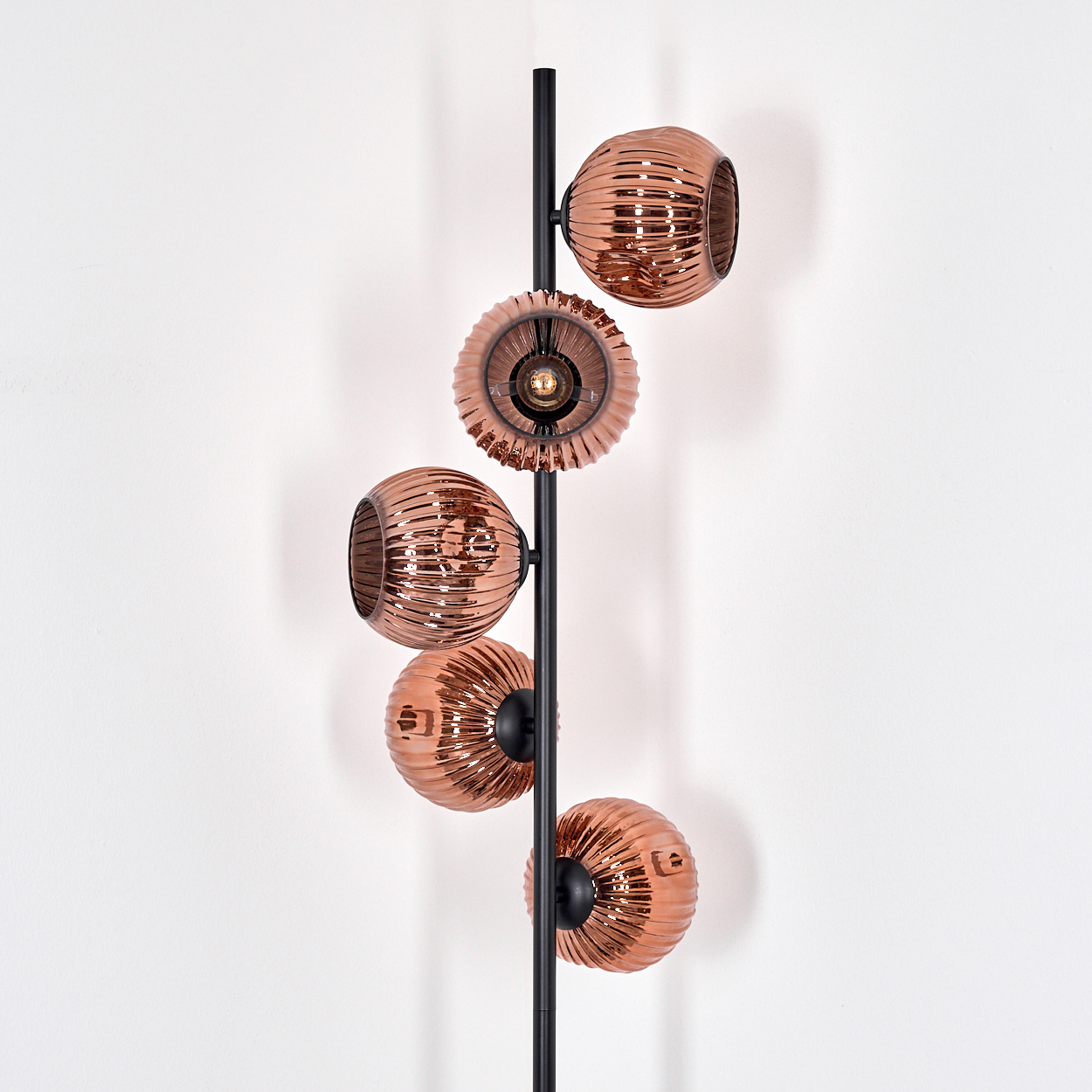 Eine moderne Stehlampe mit fünf kupferfarbenen, kugelförmigen Lampenschirmen an einer schwarzen Stange vor einer weißen Wand.