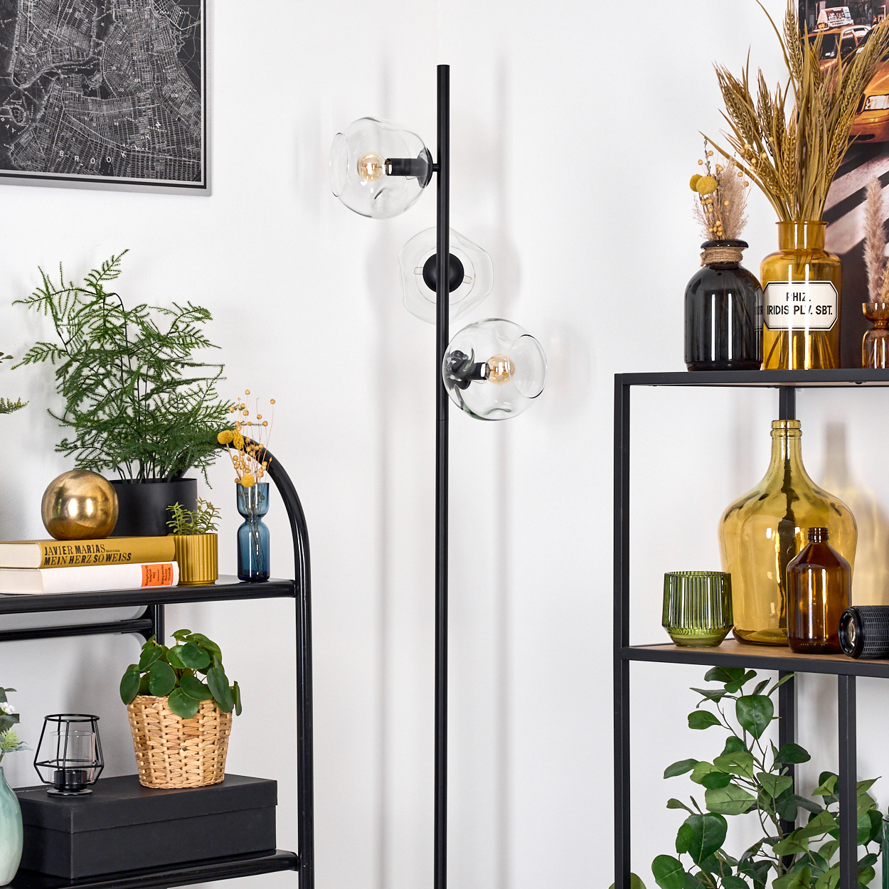 Ein Raum mit Regalen, Pflanzen und einer schwarzen Lampe mit drei Glaskugeln. Kartenkunst an der Wand.