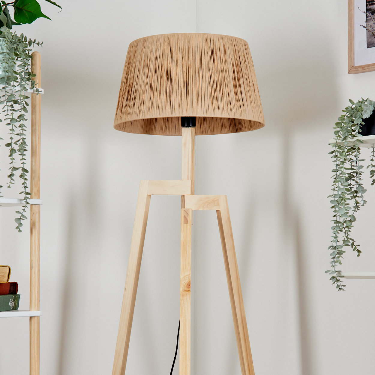 Ein beiger Lampenschirm sitzt auf einer hölzernen Stativ-Stehlampe. Im Hintergrund befinden sich grüne Pflanzen und ein Bücherregal. Die Wand ist weiß.