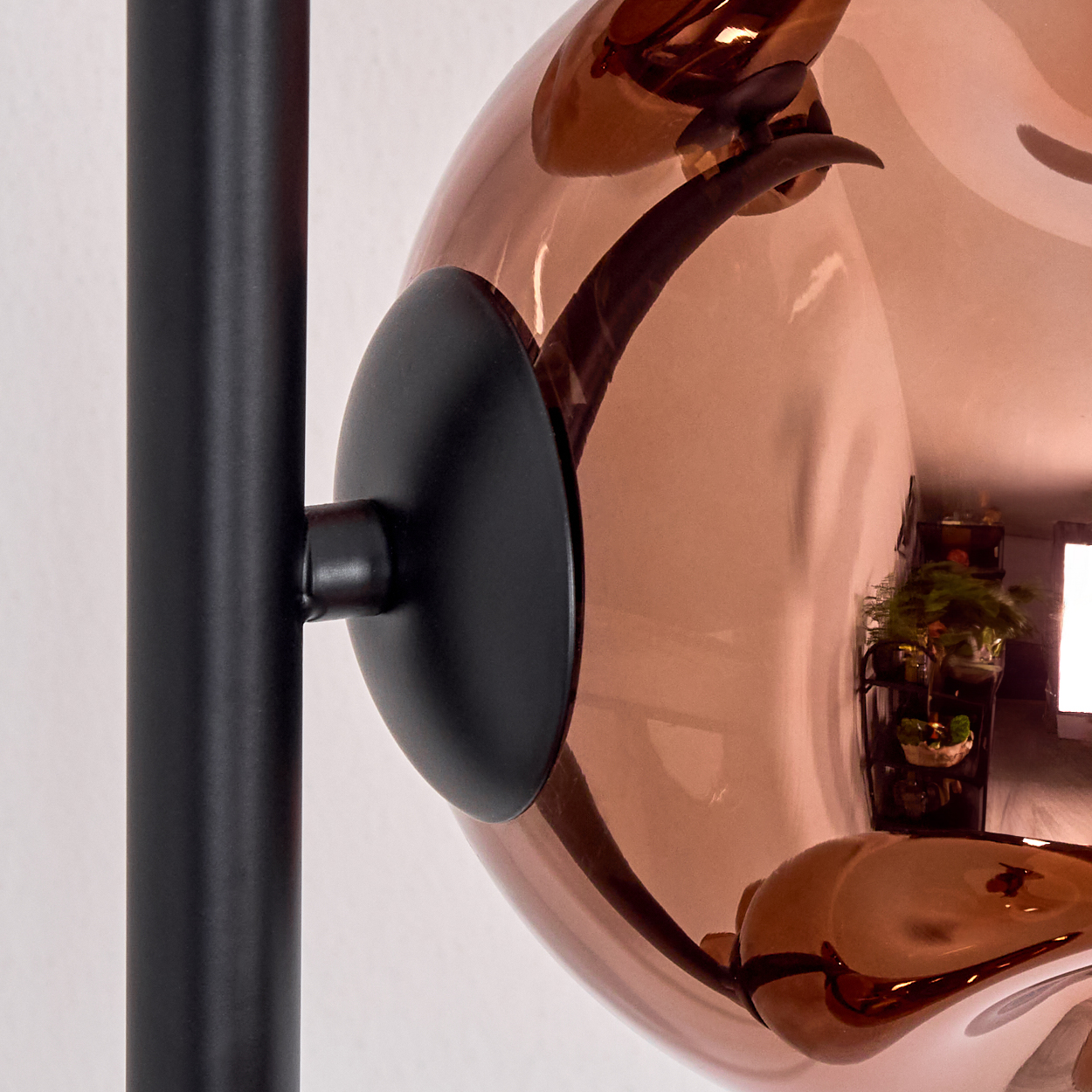 Nahaufnahme einer modernen Lampe. Eine schwarze Metallstange trägt eine kupferfarbene Glaskugel. Die Kugel reflektiert einen Raum mit Pflanzen.