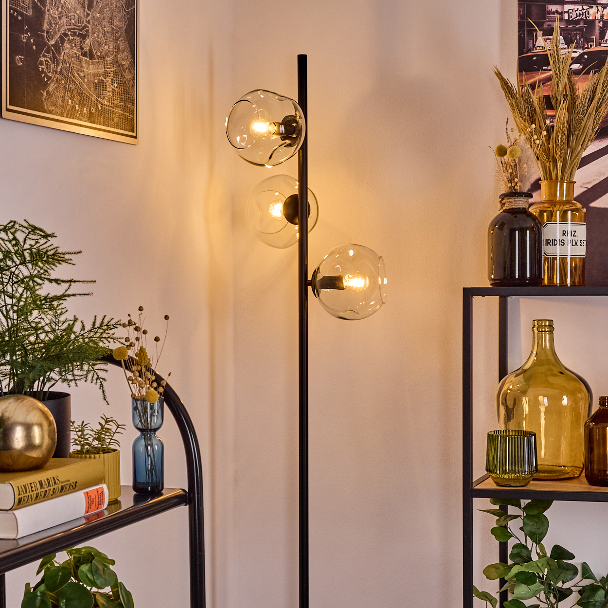 Eine moderne Stehlampe mit drei Glaskugeln. Regale halten Pflanzen, Bücher, Vasen und Dekorationsartikel. Eine Karte und ein Bild an der Wand.