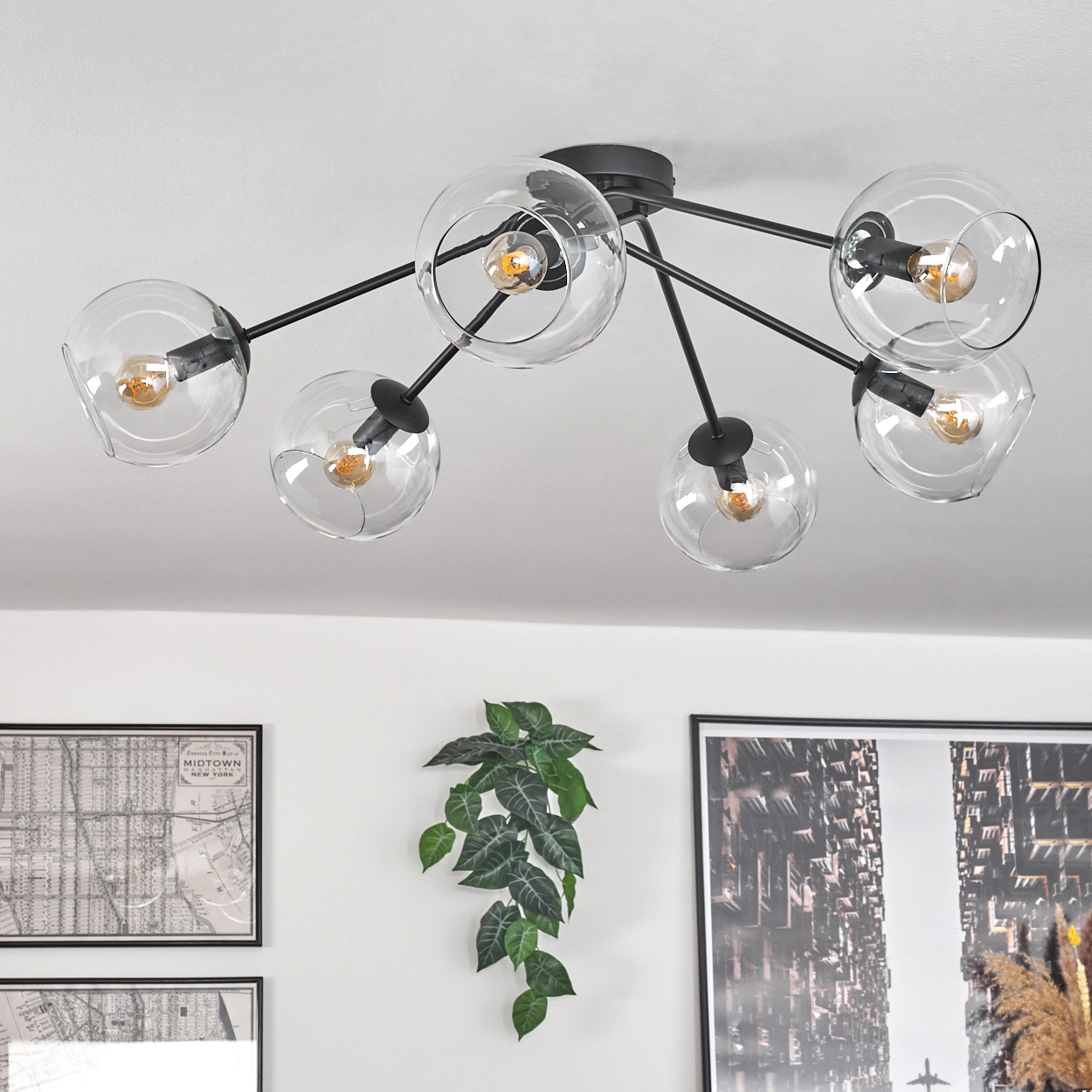 Deckenleuchte mit klaren Glaskugeln und Edison-Glühbirnen. Die Leuchte ist schwarz. Wandkunst und eine Pflanze befinden sich darunter.