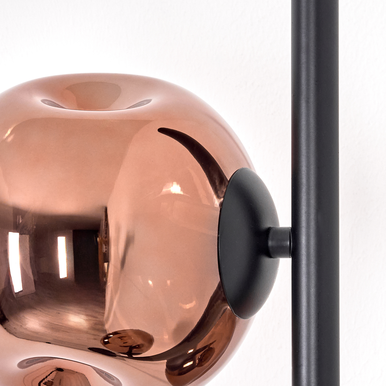 Nahaufnahme eines kupferfarbenen, kugelförmigen Lampenschirms mit schwarzer Halterung und weißer Wand im Hintergrund.
