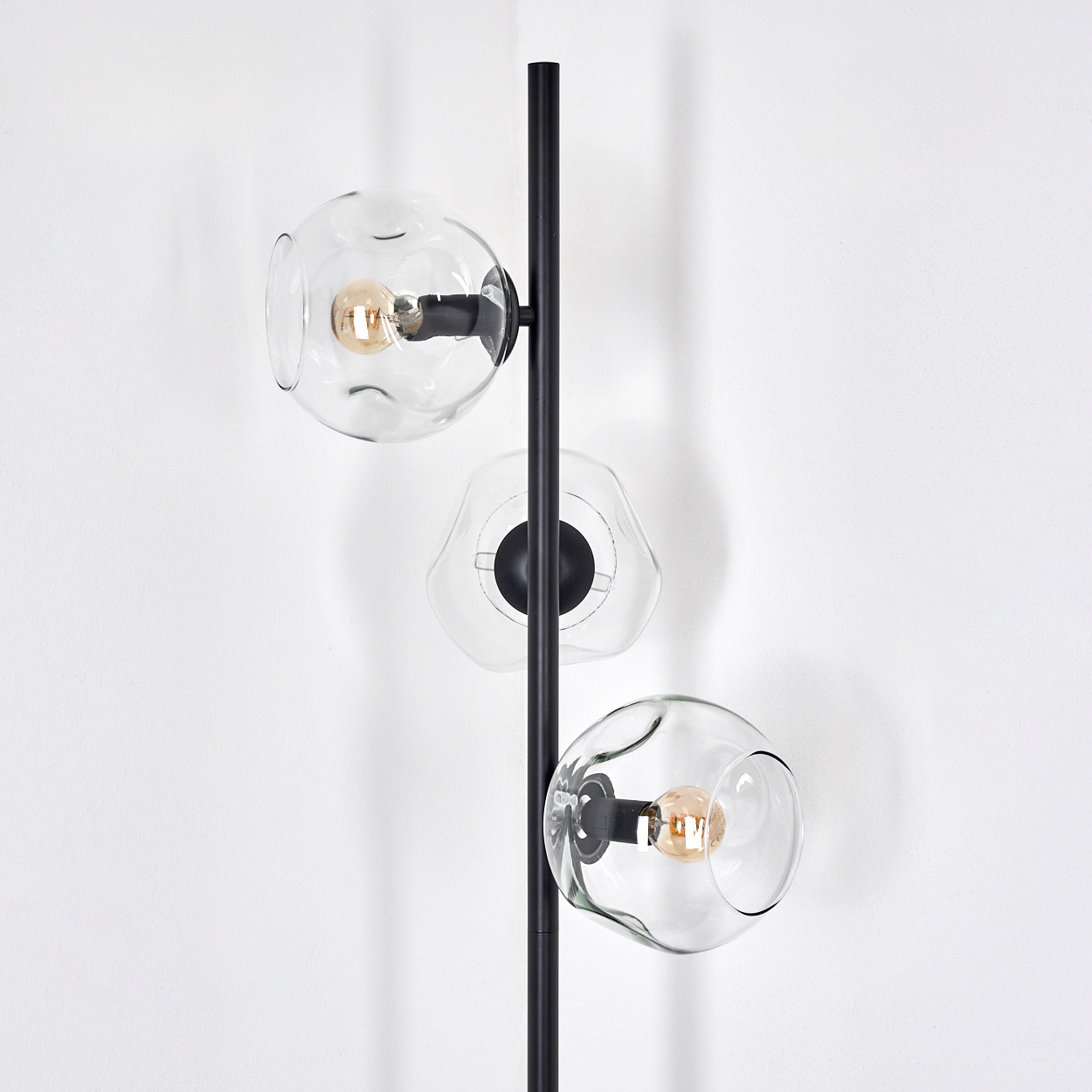 Eine moderne Stehlampe mit drei Glaskugeln, jede mit einer Glühbirne. Der Rahmen ist schwarz, vor einer weißen Wand.