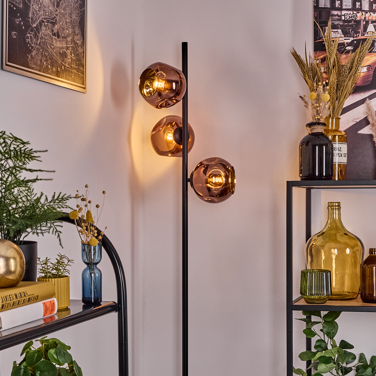 Eine moderne Stehlampe mit drei kugelförmigen, bernsteinfarbenen Glasschirmen. Bücher, Pflanzen und Dekorationsartikel stehen daneben.