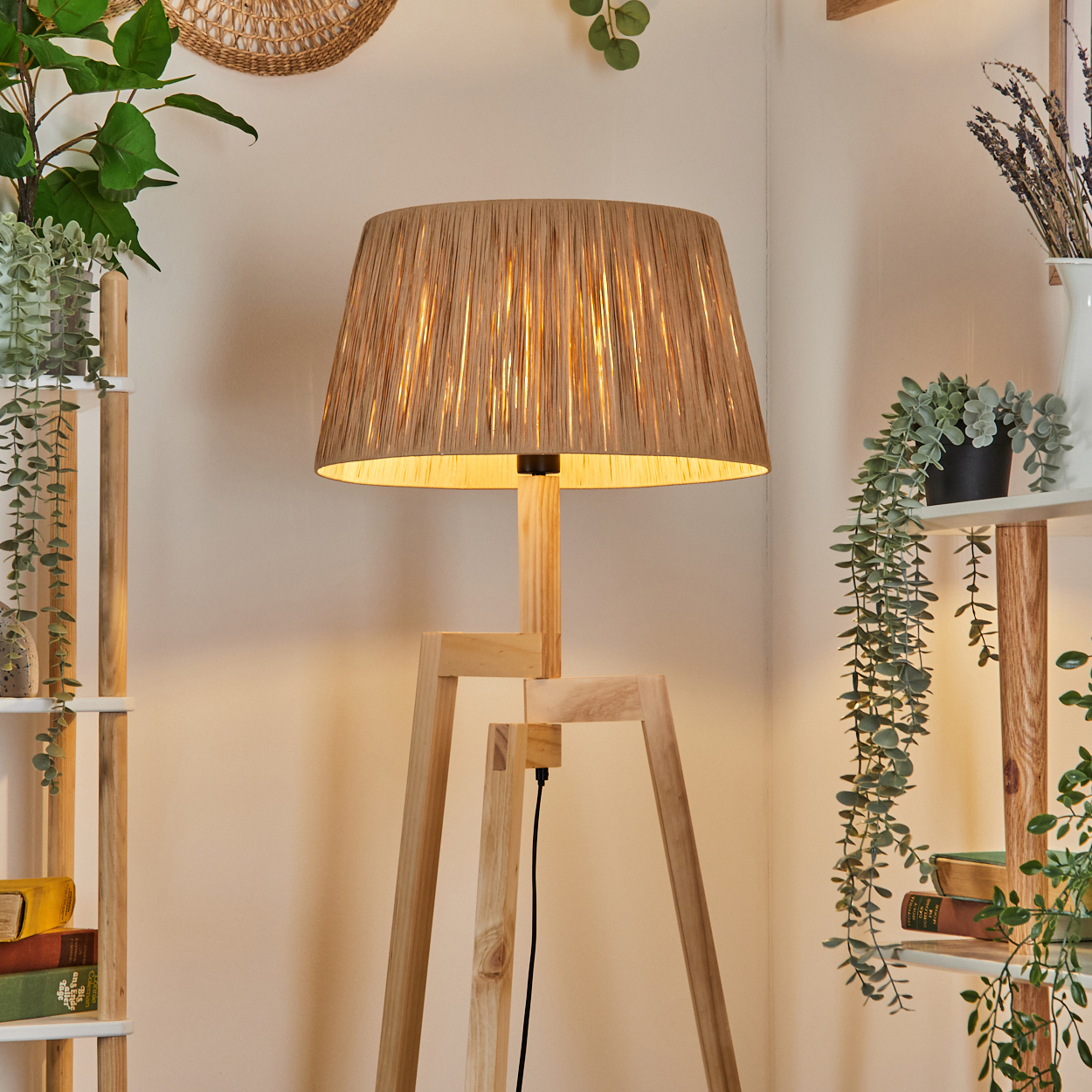 Eine Holz-Dreibein-Lampe mit beigem Lampenschirm steht in einem Raum mit Regalen und Pflanzen. Die Lampe ist beleuchtet.