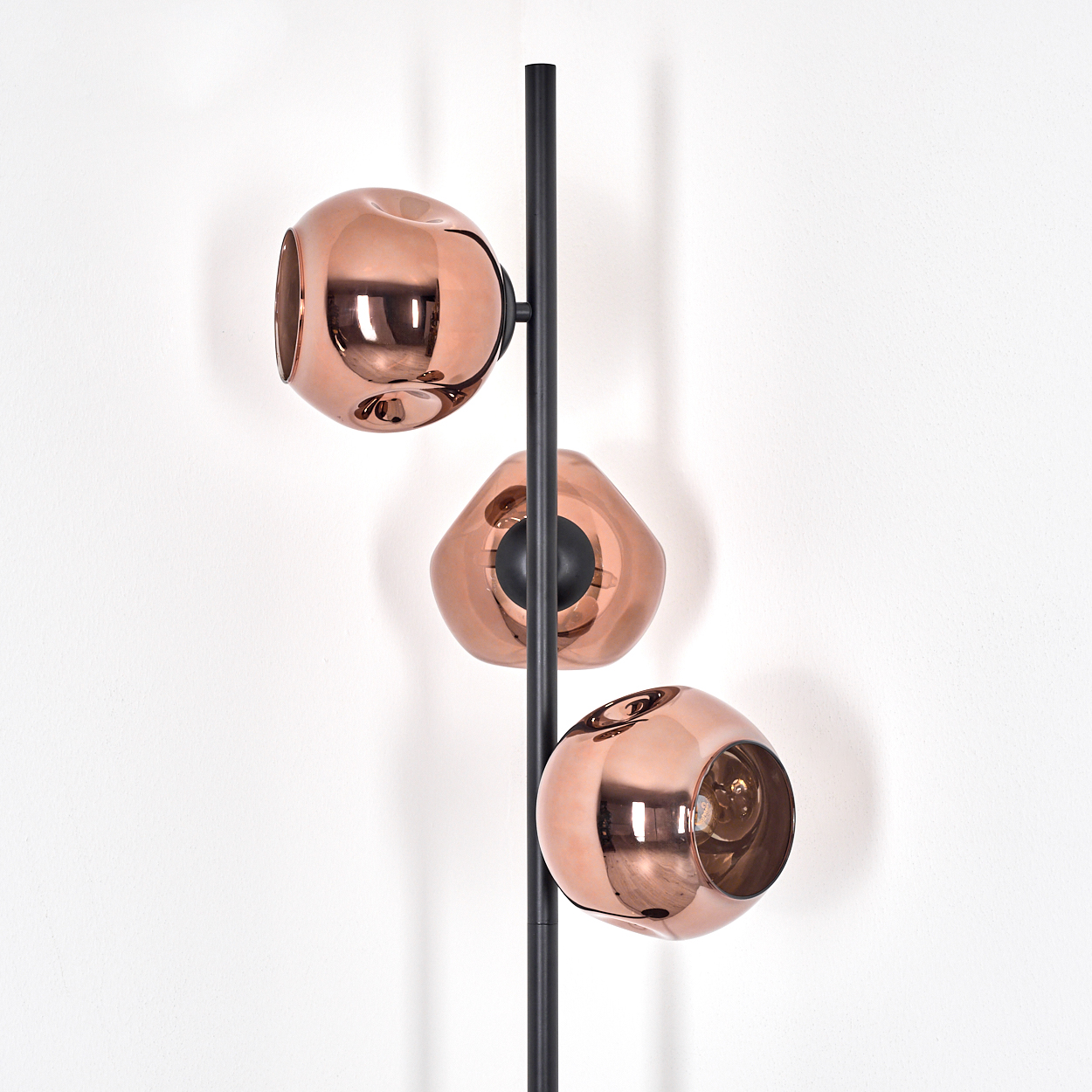 Eine moderne Lampe mit drei kupferfarbenen, kugelförmigen Schirmen vor einer weißen Wand. Sie hat eine schwarze Stange.