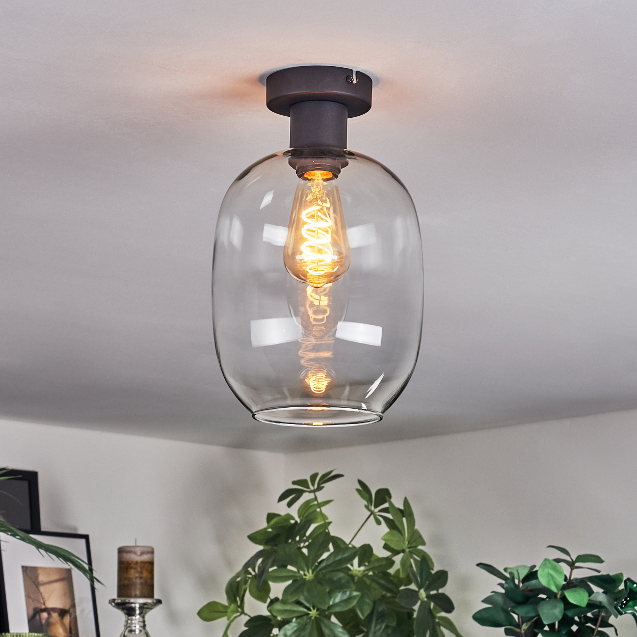Eine moderne Deckenleuchte mit Glasschirm und warmer Glühbirne. Pflanzen und eine Kerze sind im unscharfen Hintergrund zu sehen.