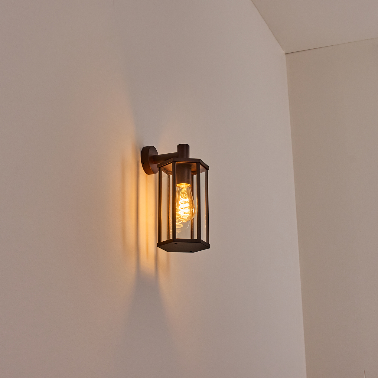 Eine dunkle, geometrische Wandlampe mit leuchtender Glühbirne an weißer Wand. Die Lampe wirft warmes Licht und Schatten.