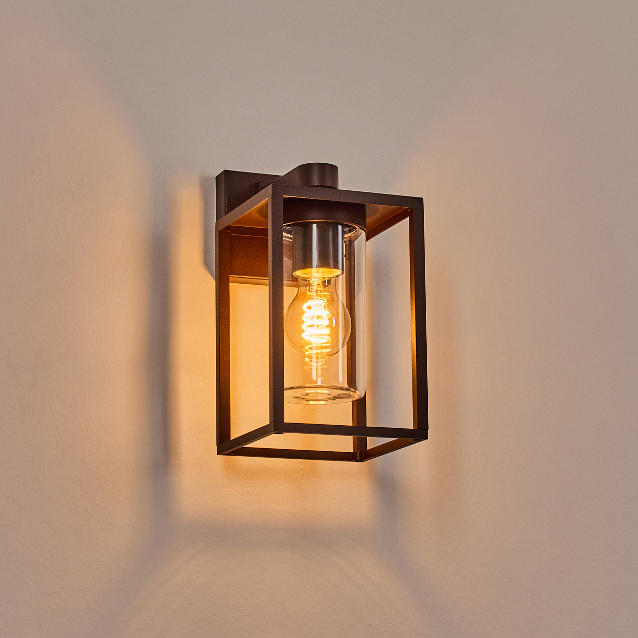 Eine dunkle Außenwandleuchte aus Metall mit einem Glaszylinder, der eine leuchtende Wendelbirne enthält, an einer cremefarbenen Wand befestigt.