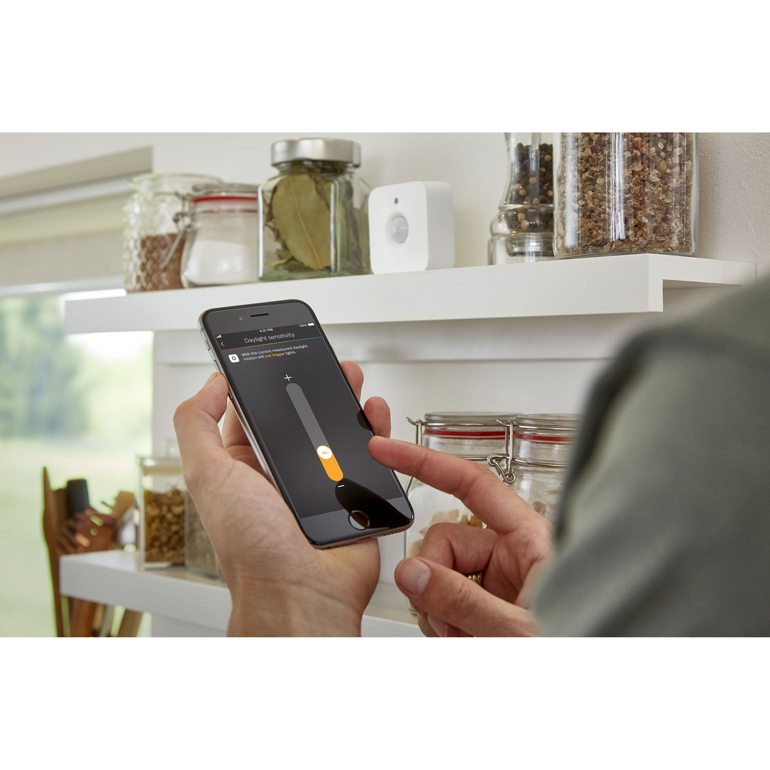 Osoba trzyma telefon, dostosowując ustawienie o nazwie 'Czułość na światło dzienne'. Słoiki z przyprawami i biały czujnik na półce.