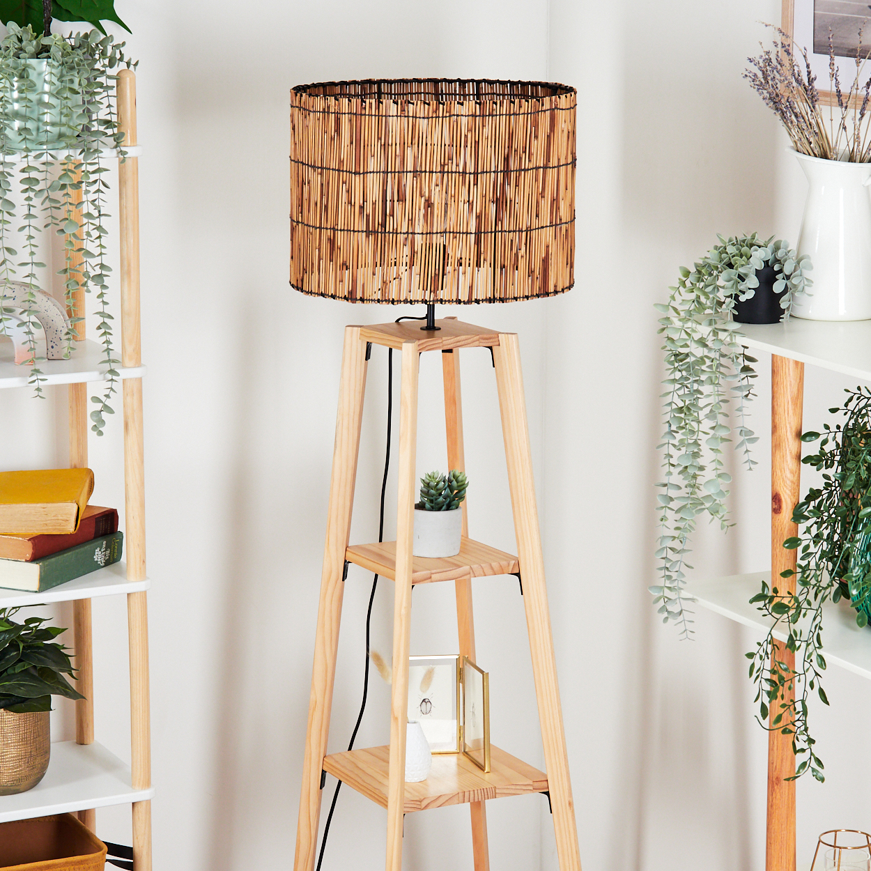 Eine Holz-Stehlampe mit gewebtem Lampenschirm steht in einem Raum mit Regalen und Pflanzen. Bücher und Dekorationsartikel sind sichtbar.