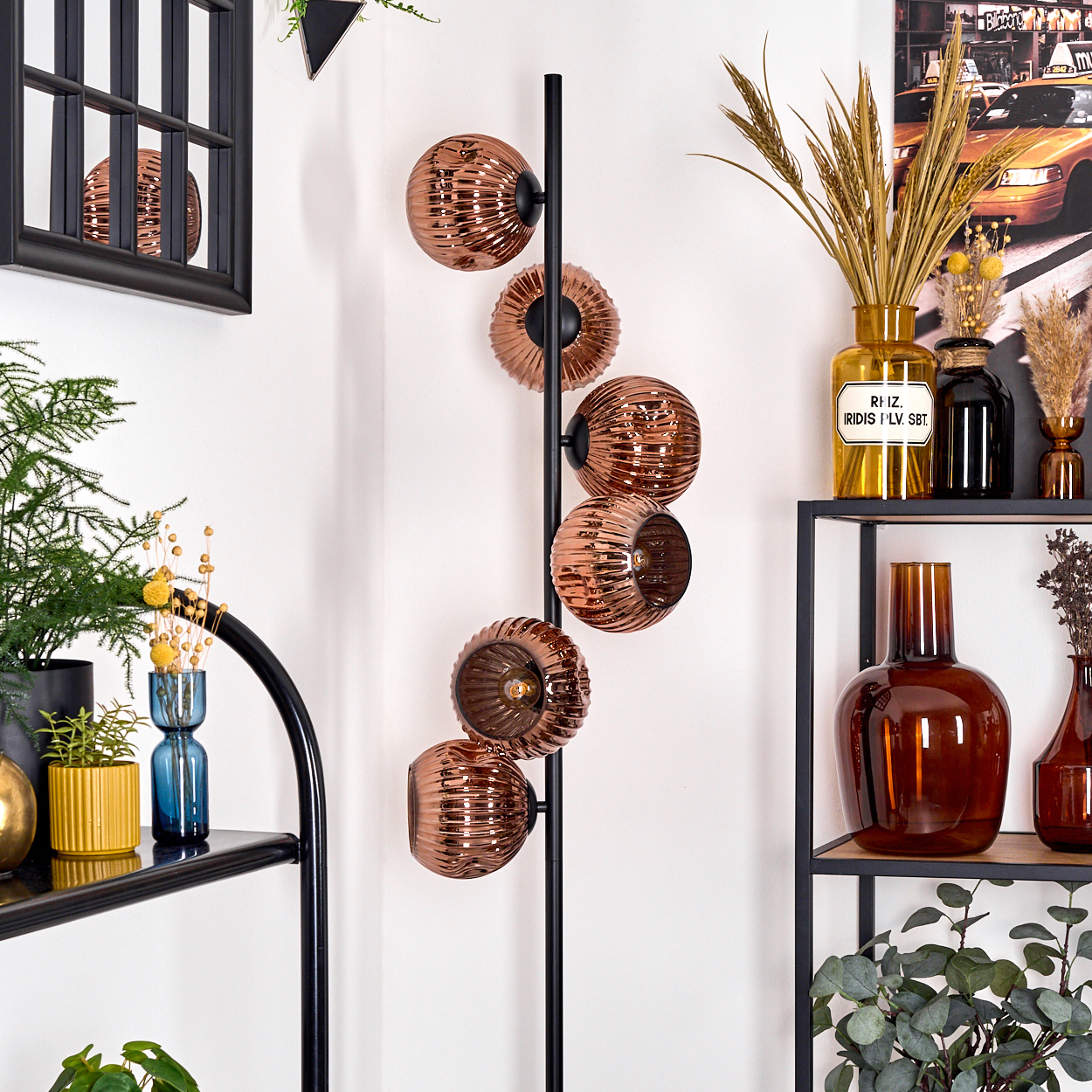 Ein modernes Interieur mit einer kupferfarbenen Stehlampe, Regalen und Vasen. Der Hintergrund ist eine weiße Wand mit Dekorationen und Pflanzen.