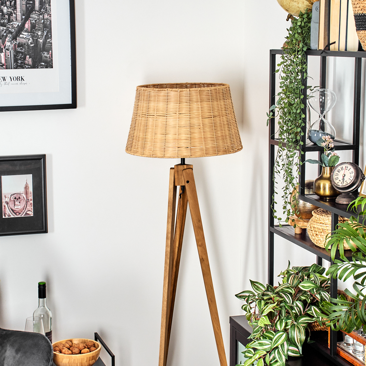 Eine gewebte Lampe steht auf Holzbeinen. Ein schwarzes Regal hat Pflanzen, eine Uhr und einen Globus. Bilder hängen an einer weißen Wand.