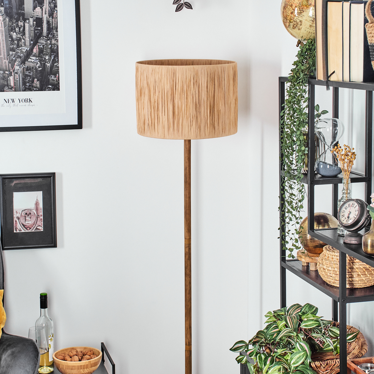 Ein Raum mit einer Stehlampe. Rechts ein Regal mit Dekorationen. Bilder links. Eine Schüssel mit Nüssen auf einem kleinen Tisch.