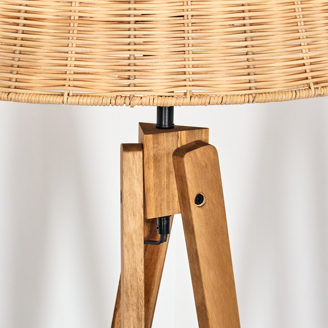 Nahaufnahme einer hölzernen Stativlampe. Der Lampenschirm ist aus geflochtenem Korb. Die Beine sind aus Holz, der Hintergrund ist eine weiße Wand.