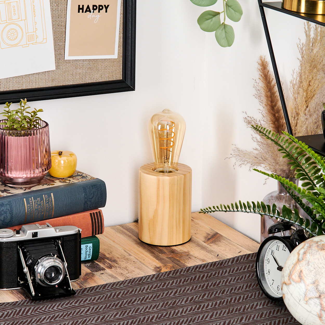 Ein Schreibtisch mit Lampe, Büchern, Kamera und Globus. Ein rosa Blumentopf, ein Apfel und ein 'Happy Day'-Schild sind sichtbar.