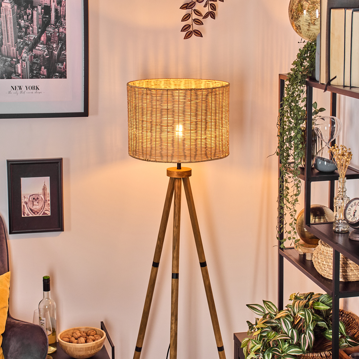 Eine gewebte Lampe steht auf Holzbeinen. Regale mit Büchern, Pflanzen und Dekoration befinden sich rechts. Bilder und eine Schüssel mit Walnüssen sind zu sehen.