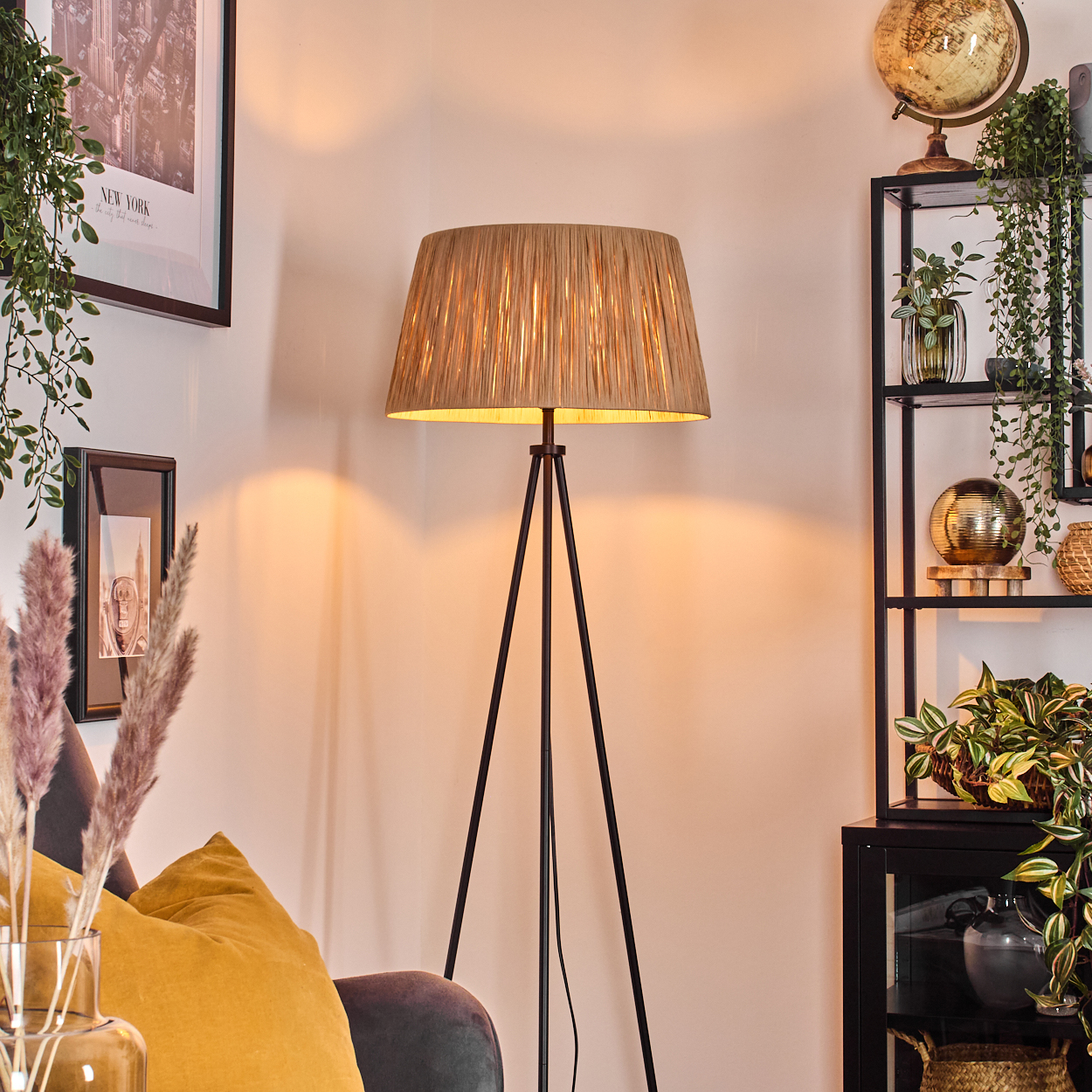 Eine beleuchtete Stehleuchte mit beigem Lampenschirm steht in einem Raum mit Regal und Sofa, Pflanzen und Globus.