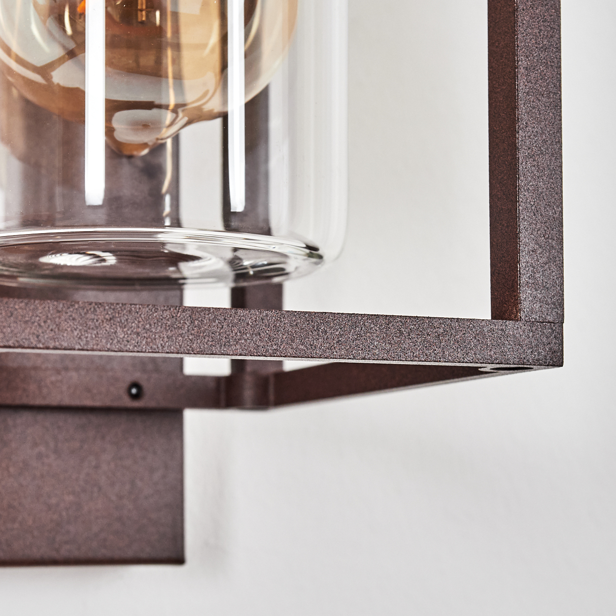 Nahaufnahme einer braunen Metallrahmen-Leuchte mit einem Glaszylinder, der eine Glühbirne umschließt, vor einer weißen Wand.