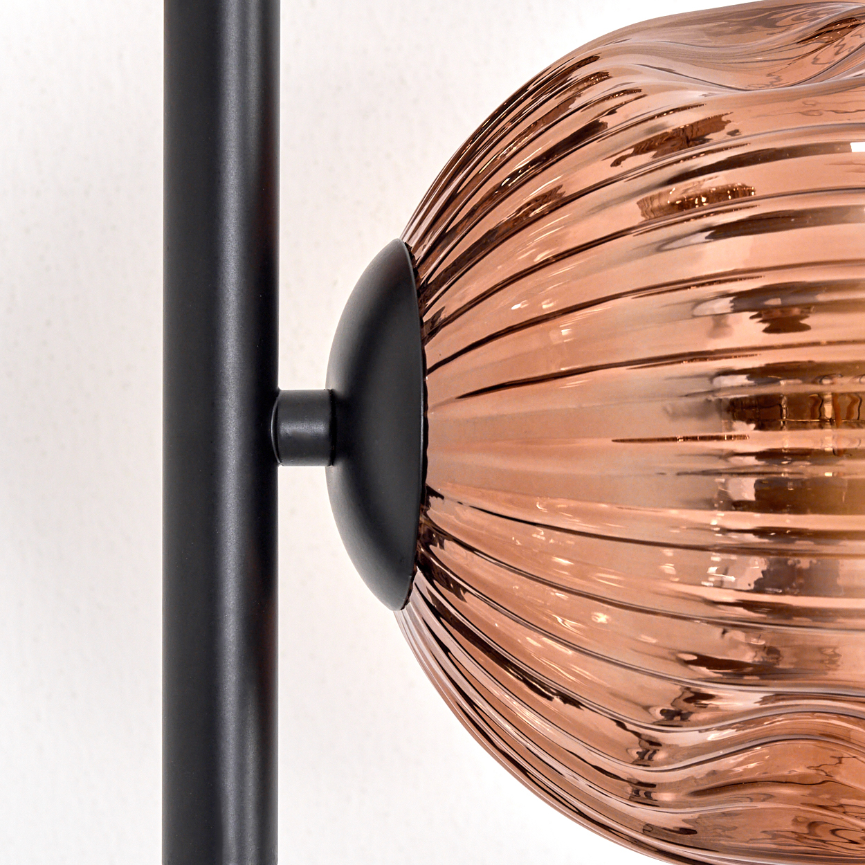 Nahaufnahme einer Lampe. Eine schwarze Metallstange trägt eine kupferfarbene, strukturierte Glaskugel vor einer weißen Wand.