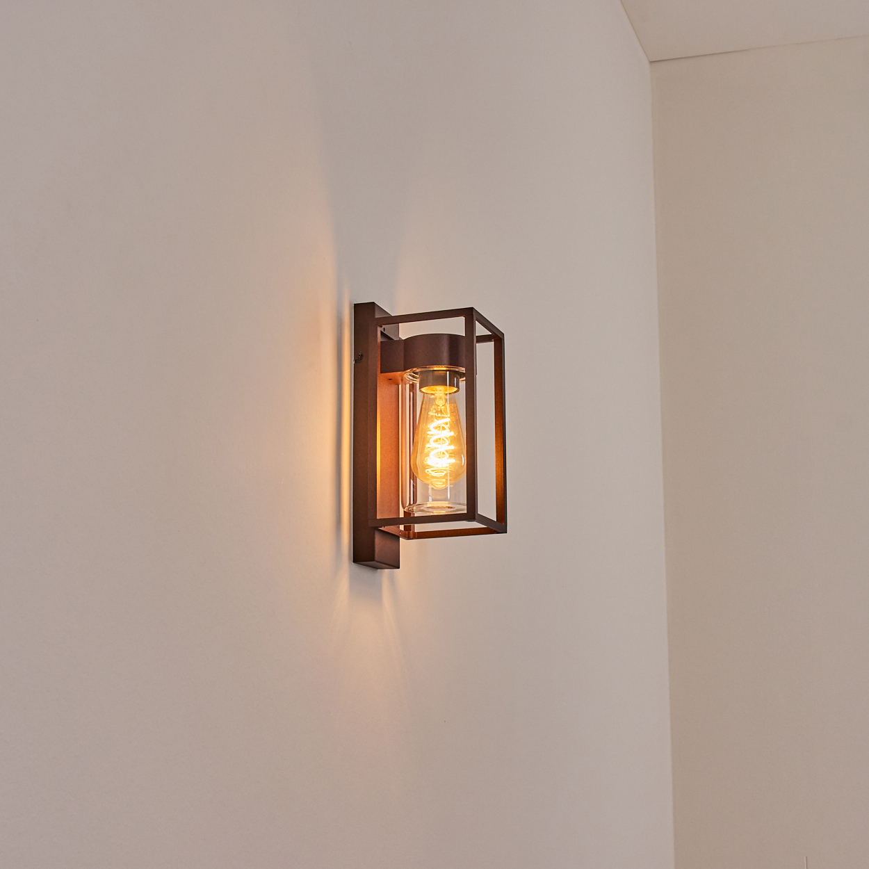 Eine dunkle Metall-Wandleuchte mit einem durchsichtigen Glaszylinder und einer leuchtenden Edison-Glühbirne an einer weißen Wand.