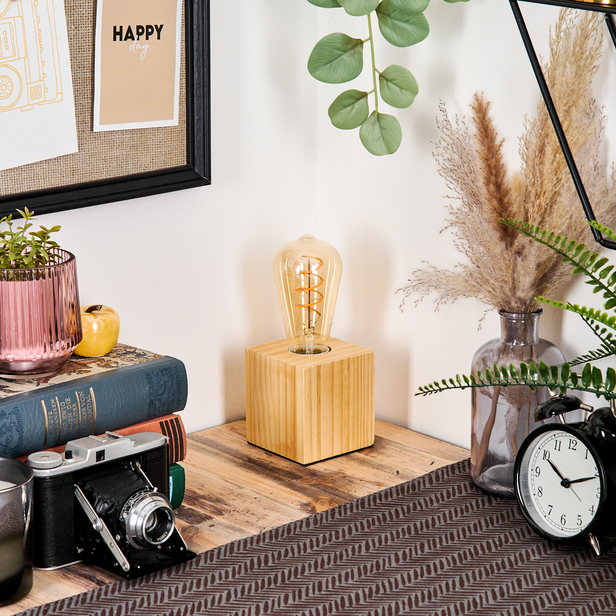 Ein Schreibtisch mit Lampe, Büchern, Kamera, Uhr und Dekorationen. Ein 'Happy day'-Hinweis an der Wand. Die Szene hat eine warme, gemütliche Atmosphäre.