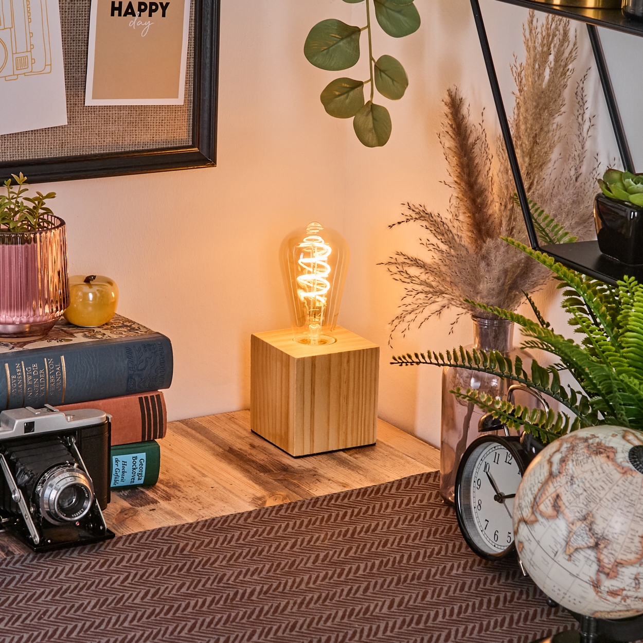 Ein Schreibtisch mit einer beleuchteten Glühbirne, Büchern, Kamera, Uhr und Globus. Dekorative Pflanzen im Hintergrund mit einem 'Happy Day' Zettel.