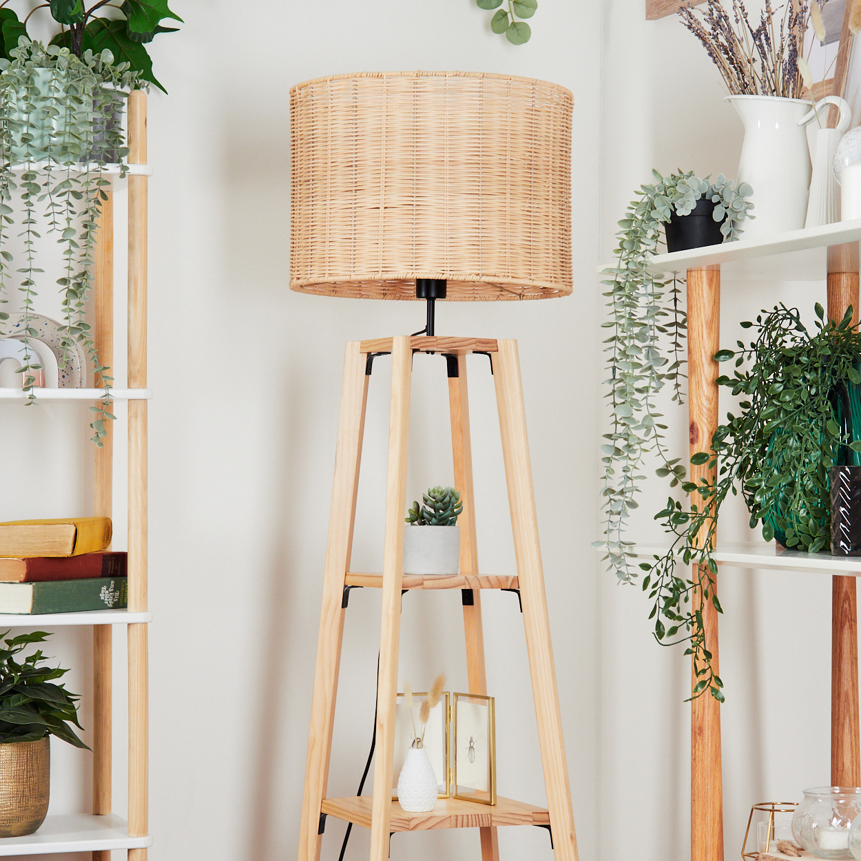 Eine gewebte Lampe steht auf einem Holzständer. Regale auf beiden Seiten halten Pflanzen, Bücher und Dekorationsartikel. Der Hintergrund ist eine weiße Wand.
