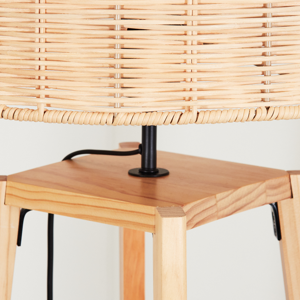 Nahaufnahme einer Lampe. Die Lampe hat einen gewebten, hellbraunen Lampenschirm, einen schwarzen Ständer und einen Holzsockel mit Beinen. Weißer Hintergrund.