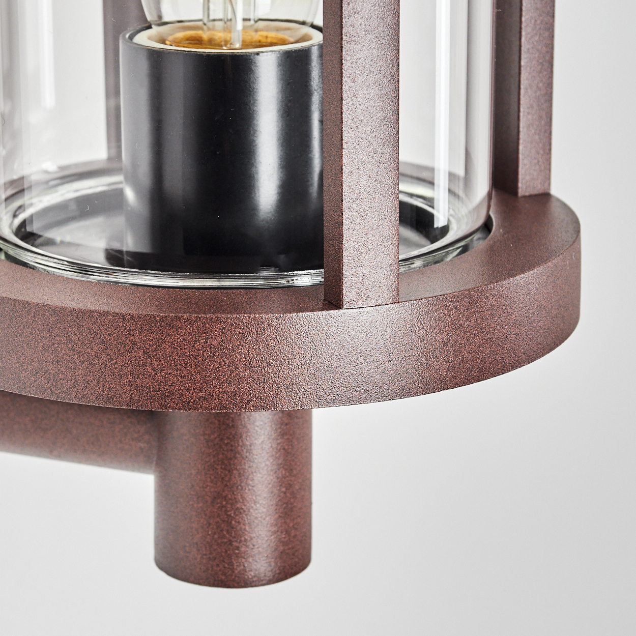 Nahaufnahme einer bronzefarbenen Außenleuchte. Sie hat einen Glaszylinder um einen schwarzen Glühbirnenhalter. Der Hintergrund ist weiß.