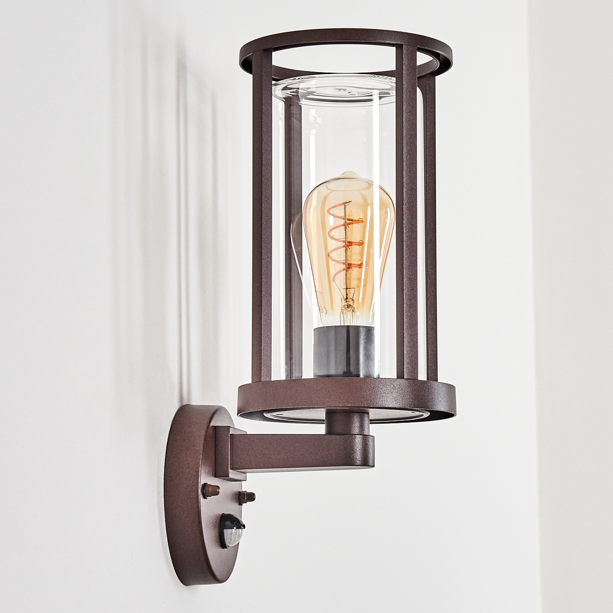 Eine bronzefarbene Wandlampe mit einem durchsichtigen Glaszylinder. Im Inneren befindet sich eine glühende Glühbirne mit Spiralfaden. Die Lampe ist an einer weißen Wand befestigt.