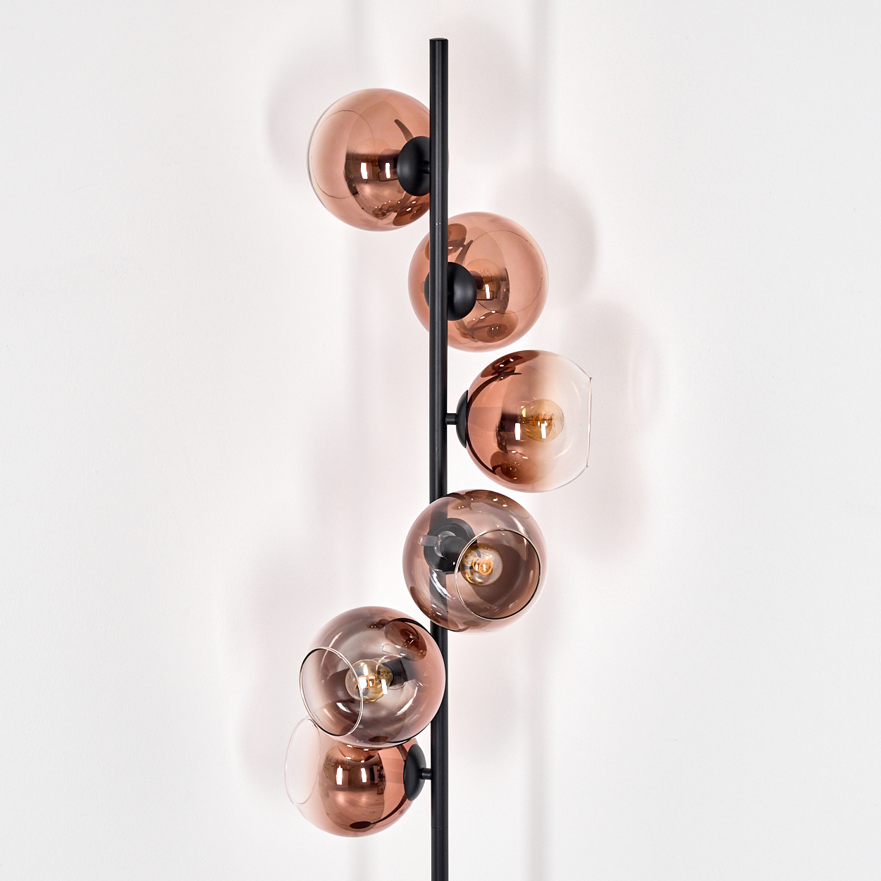 Eine moderne Lampe mit sechs kupferfarbenen Glaskugelschirmen, die an einer schwarzen vertikalen Stange befestigt sind, vor einer weißen Wand.