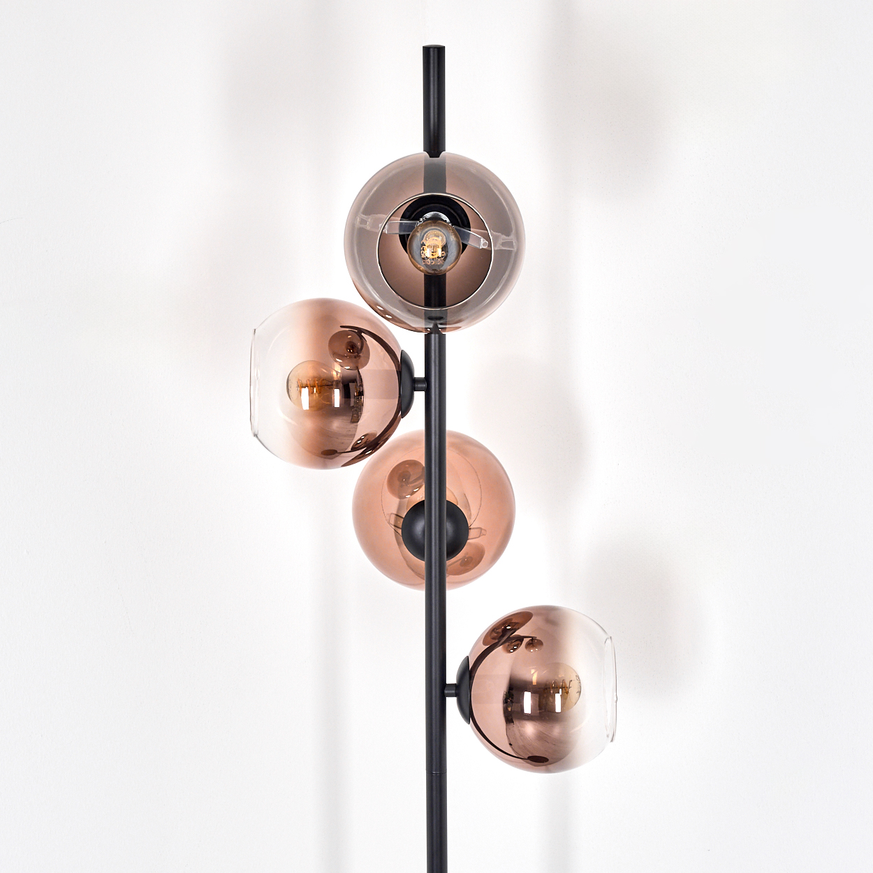 Eine moderne Stehlampe mit vier kupferfarbenen Glaskugelschirmen. Die Lampe hat ein schwarzes Metallgestell vor einer weißen Wand.