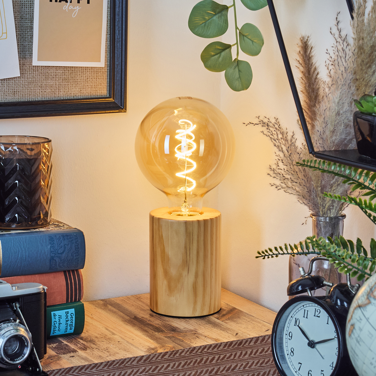 Eine beleuchtete Glühbirnenlampe auf einem Holzsockel steht auf einem Tisch. Bücher, eine Kamera und eine Uhr stehen auf dem Tisch.
