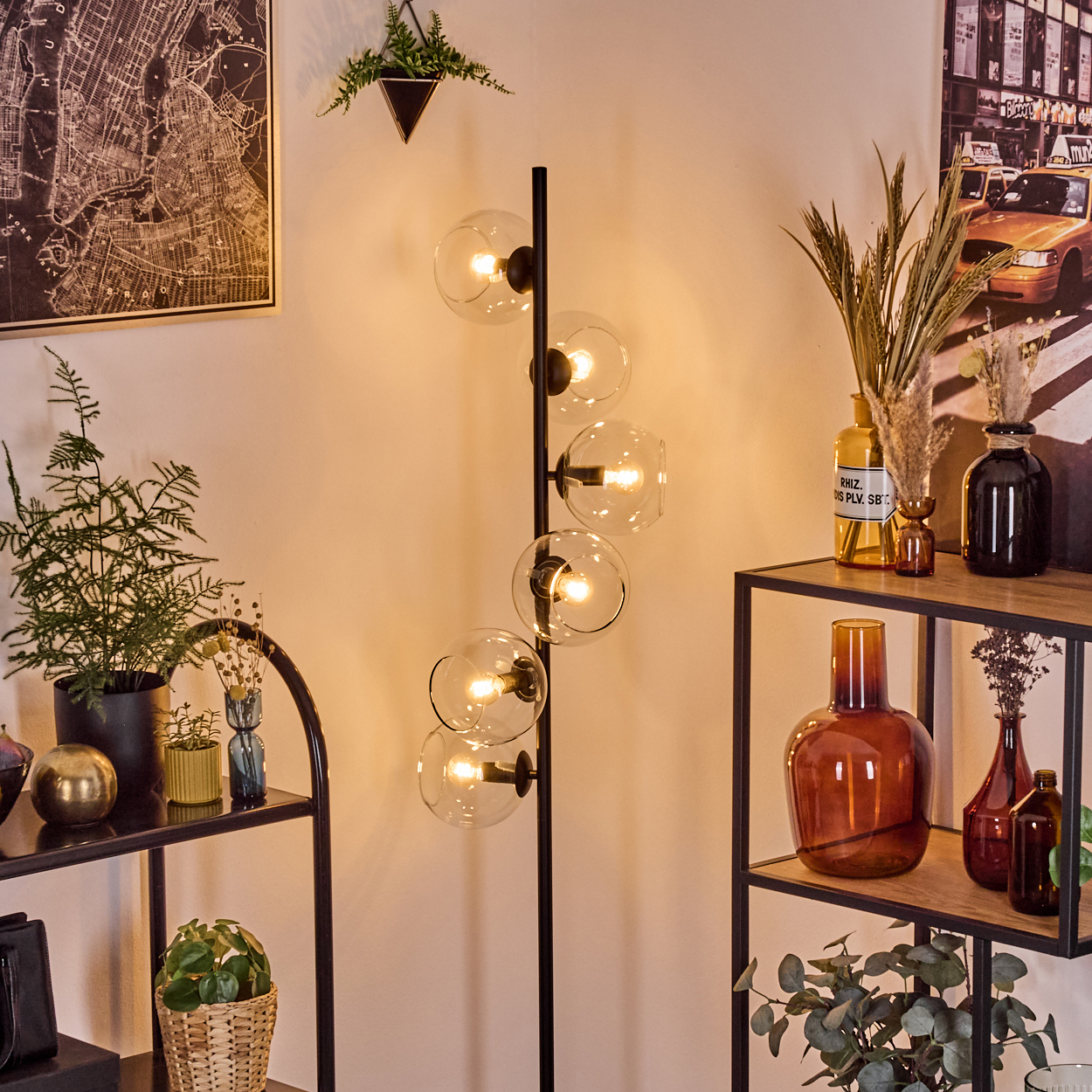Ein modernes Zimmer mit einer schwarzen Stehlampe mit Glaskugeln. Regale halten Pflanzen, Vasen. Eine Karte und ein Taxibild hängen an den Wänden.