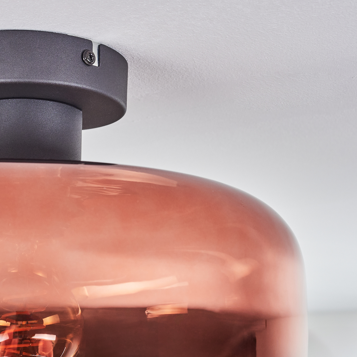 Nahaufnahme einer Deckenlampe. Sie hat einen runden, kupferfarbenen Glasschirm und einen dunkelgrauen Metallsockel. Die Decke ist weiß.