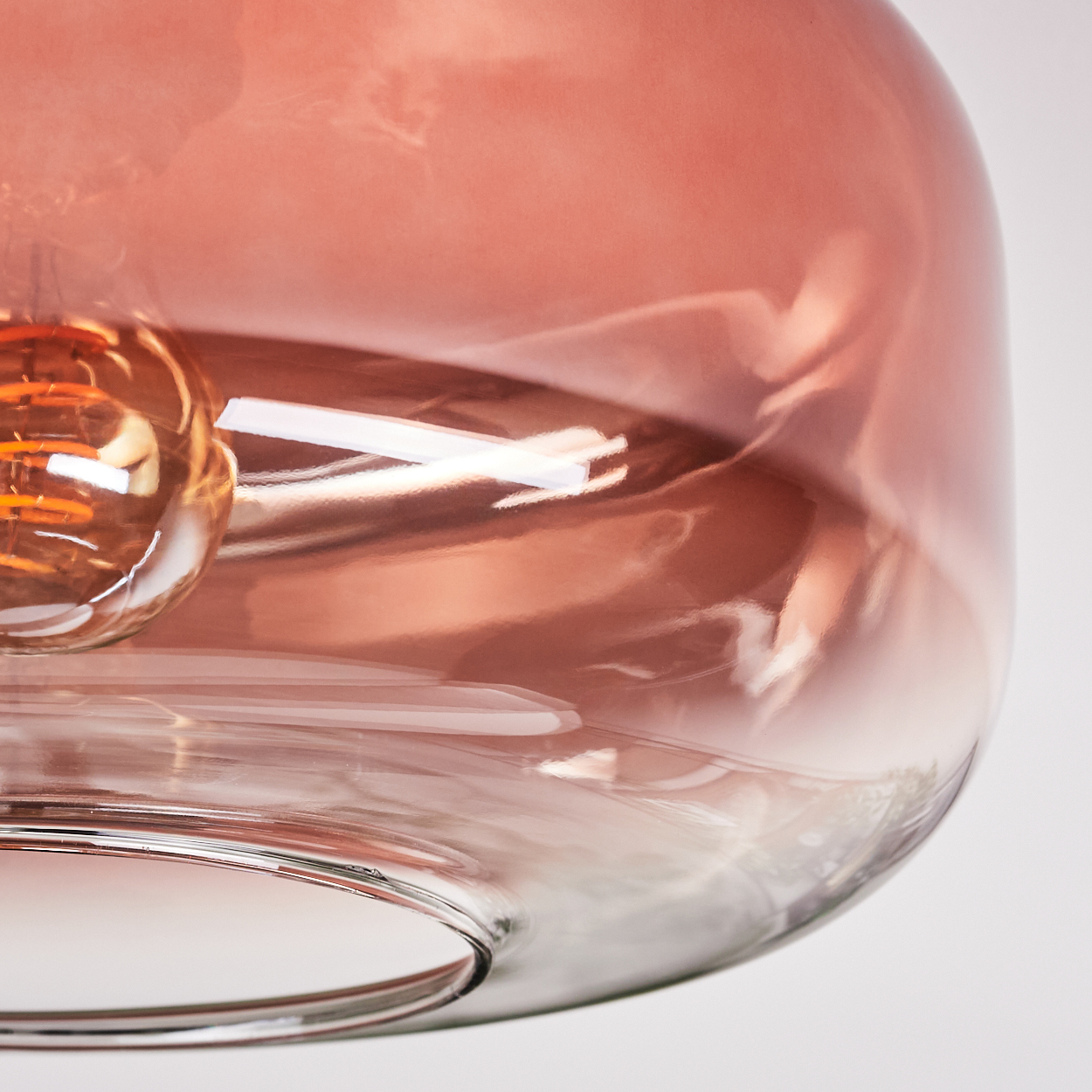 Nahaufnahme einer Glas-Pendelleuchte. Das Glas ist rosafarben mit einer Glühbirne im Inneren. Der Hintergrund ist weiß.