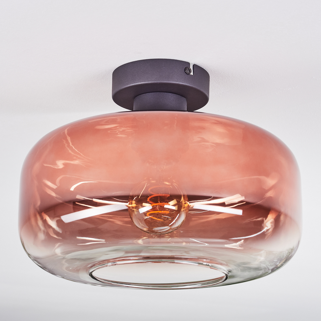 Eine moderne Deckenlampe mit rosa Glasschirm und sichtbarer Glühbirne. Sie ist an einer dunkelgrauen Halterung an der weißen Decke befestigt.