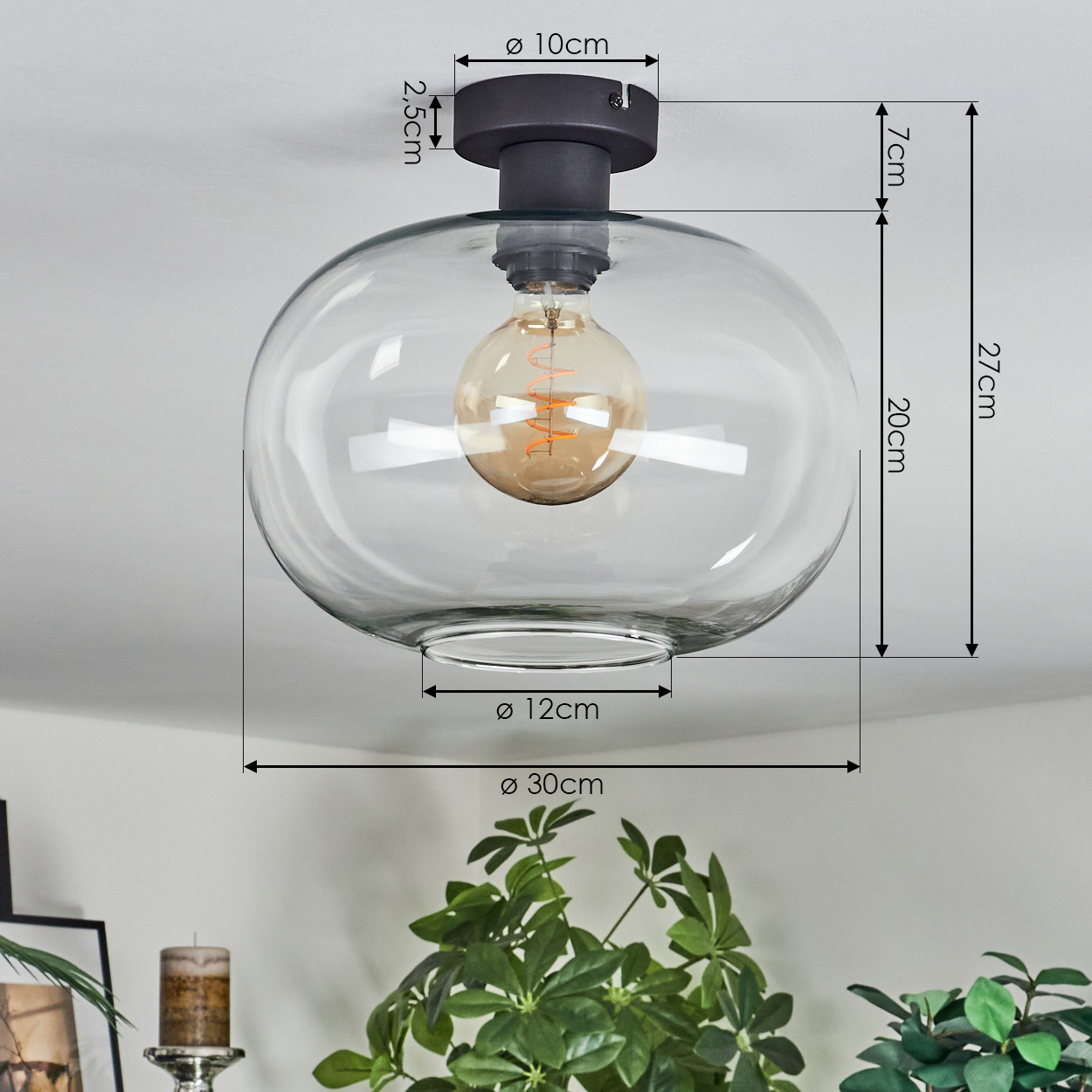 Eine klare Deckenlampe mit sichtbarer Glühbirne. Abmessungen sind angegeben. Grüne Pflanzen im Vordergrund.