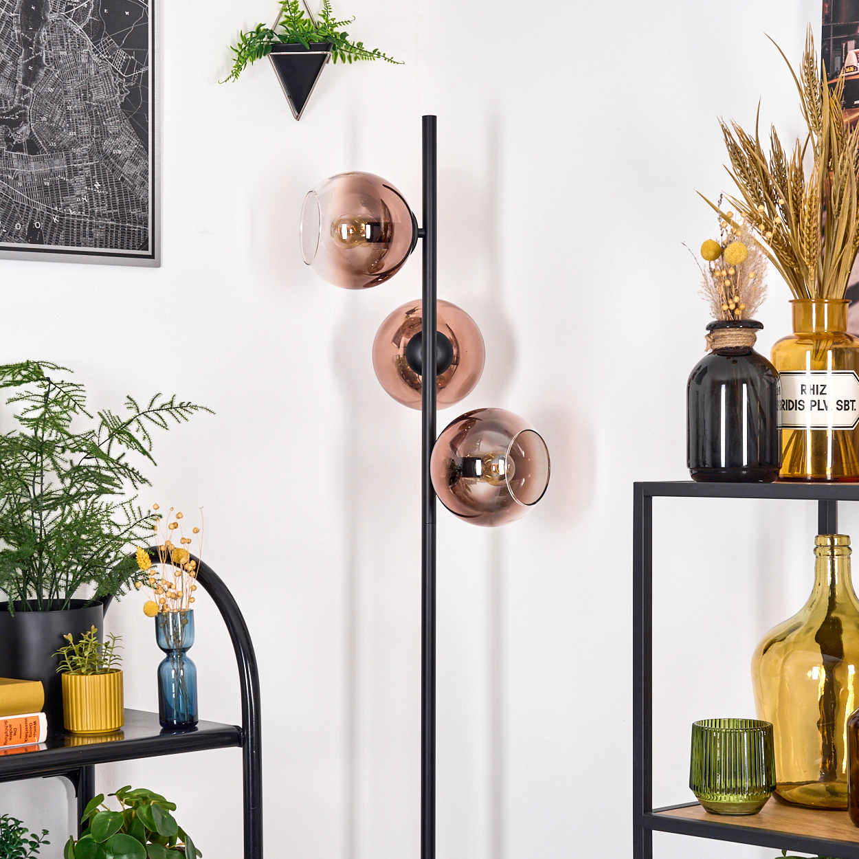 Ein modernes Zimmer mit einer schwarzen Lampe mit drei kupferfarbenen Glasschirmen. Regale zeigen Vasen, Pflanzen und eine Karte.