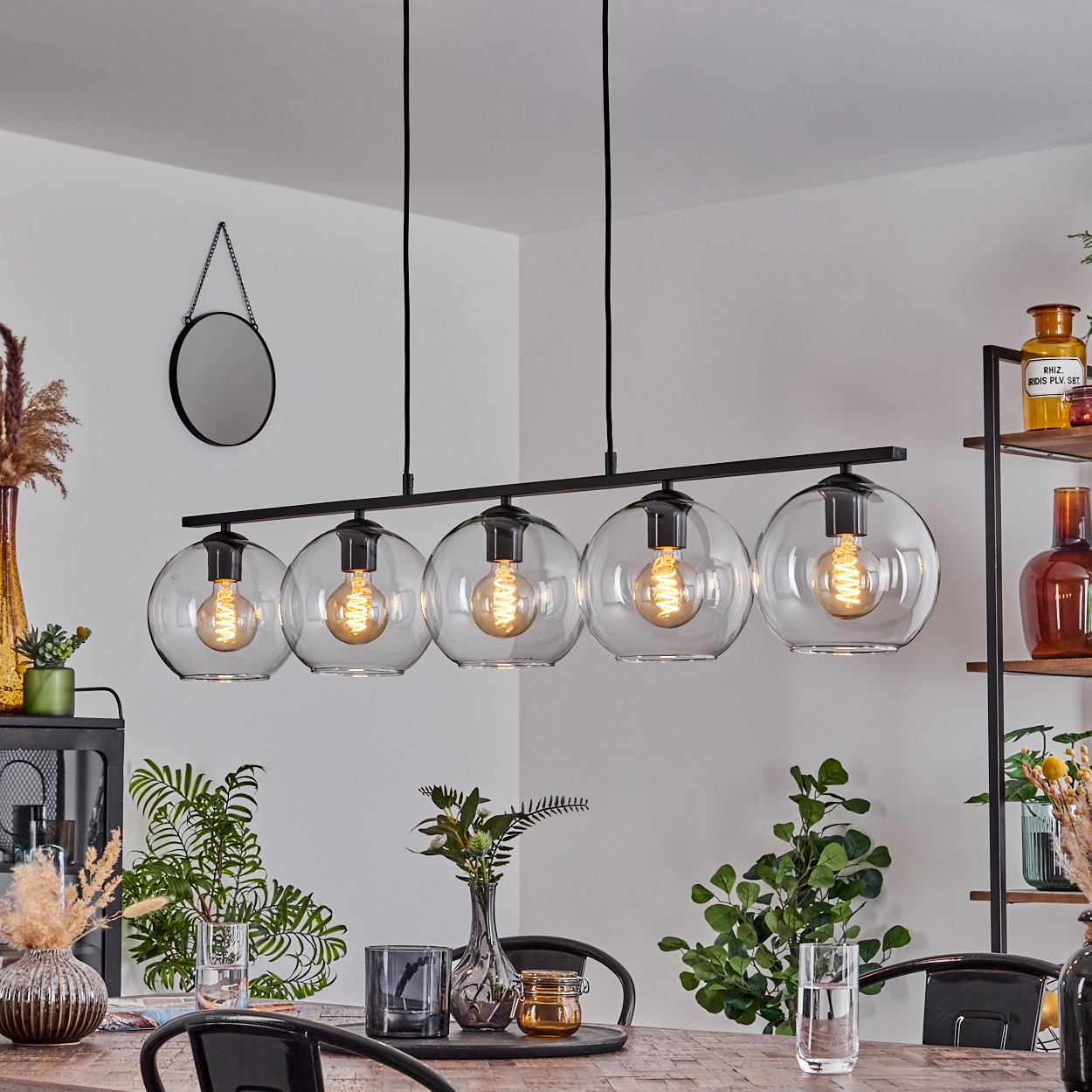 Esszimmer mit schwarzem Kronleuchter mit fünf Glaskugeln. Verschiedene Pflanzen, ein Spiegel und ein Holztisch sind vorhanden.